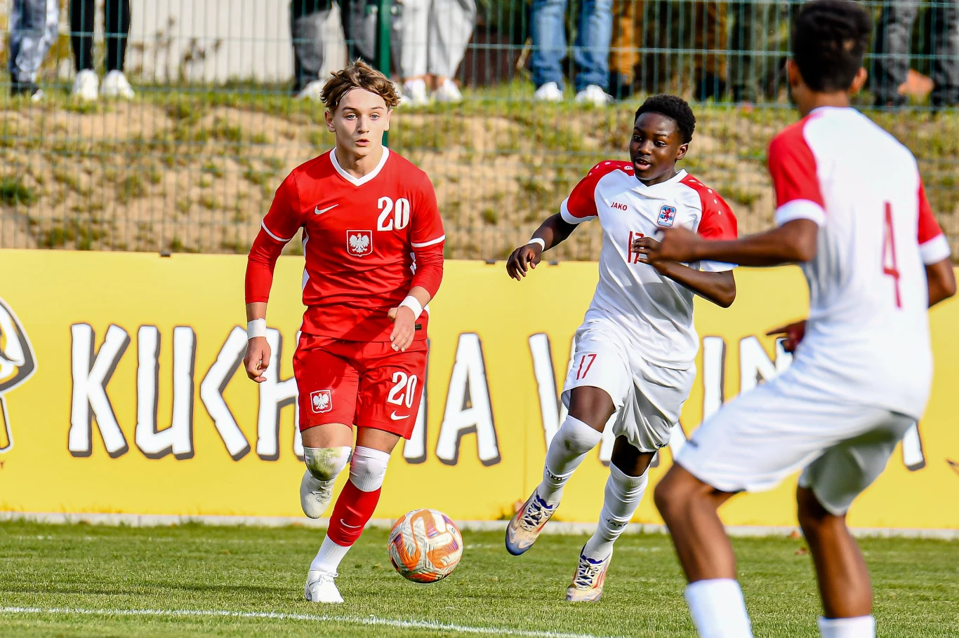 Zawodnicy w czerwono-białych strojach biegną za piłką na zielonym boisku, w tle widoczny baner reklamowy i ogrodzenie.