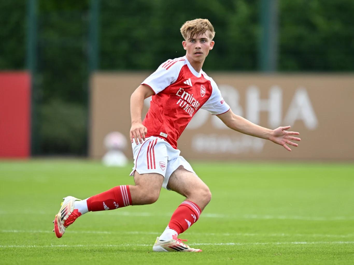 Młody piłkarz w stroju drużyny Arsenal w trakcie gry na zielonym boisku, skupiony na piłce, dynamiczna pozycja świadcząca o aktywności sportowej.