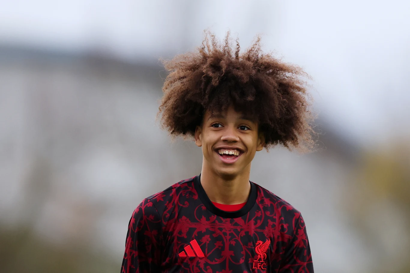 Młody piłkarz ubrany w czarno-czerwony sportowy strój z logo Liverpool FC, uśmiechnięty z charakterystyczną bujną fryzurą afro, znajduje się na tle rozmytego krajobrazu.