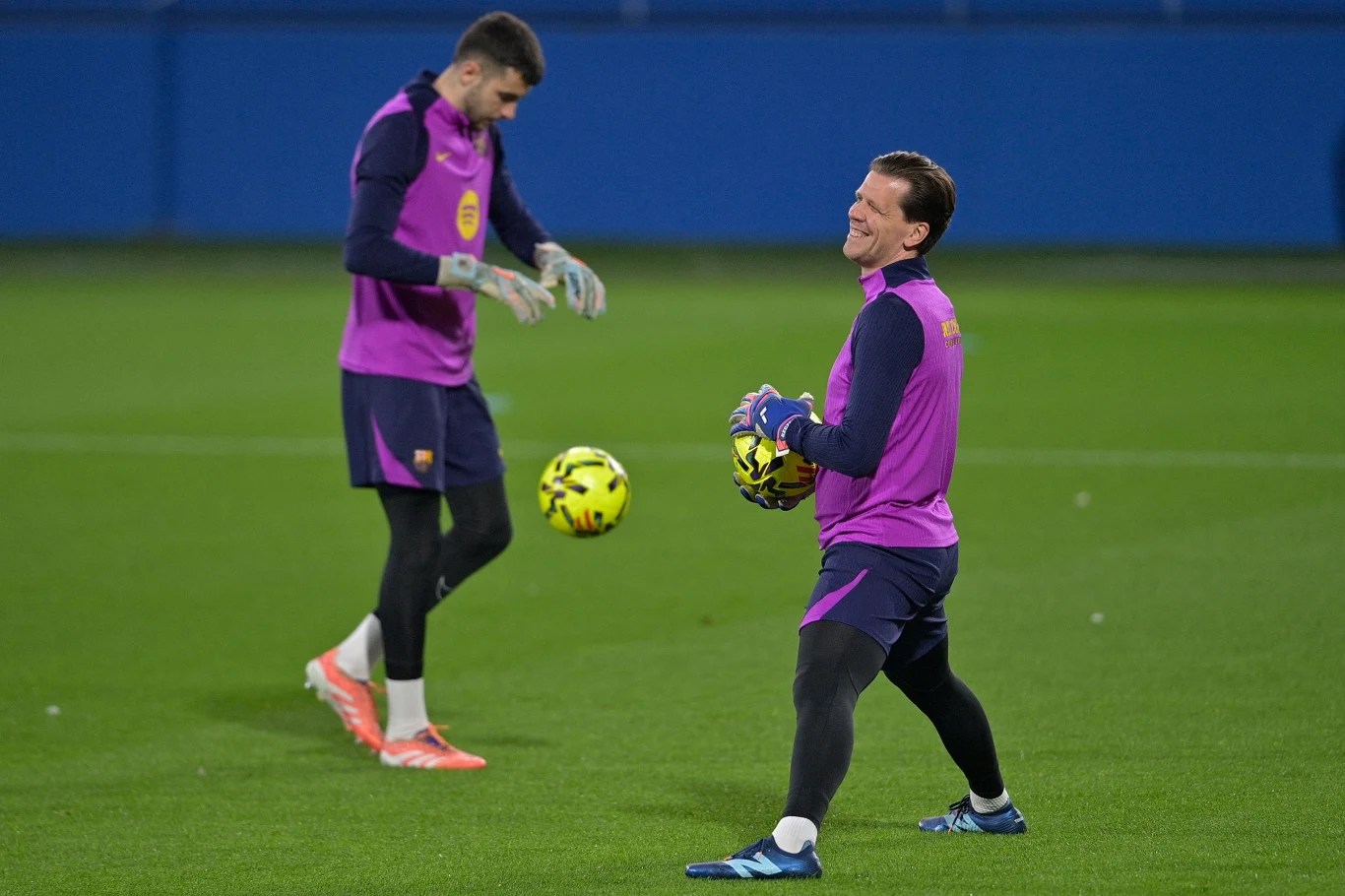 Dwóch piłkarzy ubranych w fioletowe stroje treningowe na zielonym boisku piłkarskim, jeden z nich trzyma piłkę, drugi przygotowuje się do ćwiczenia bramkarskiego.