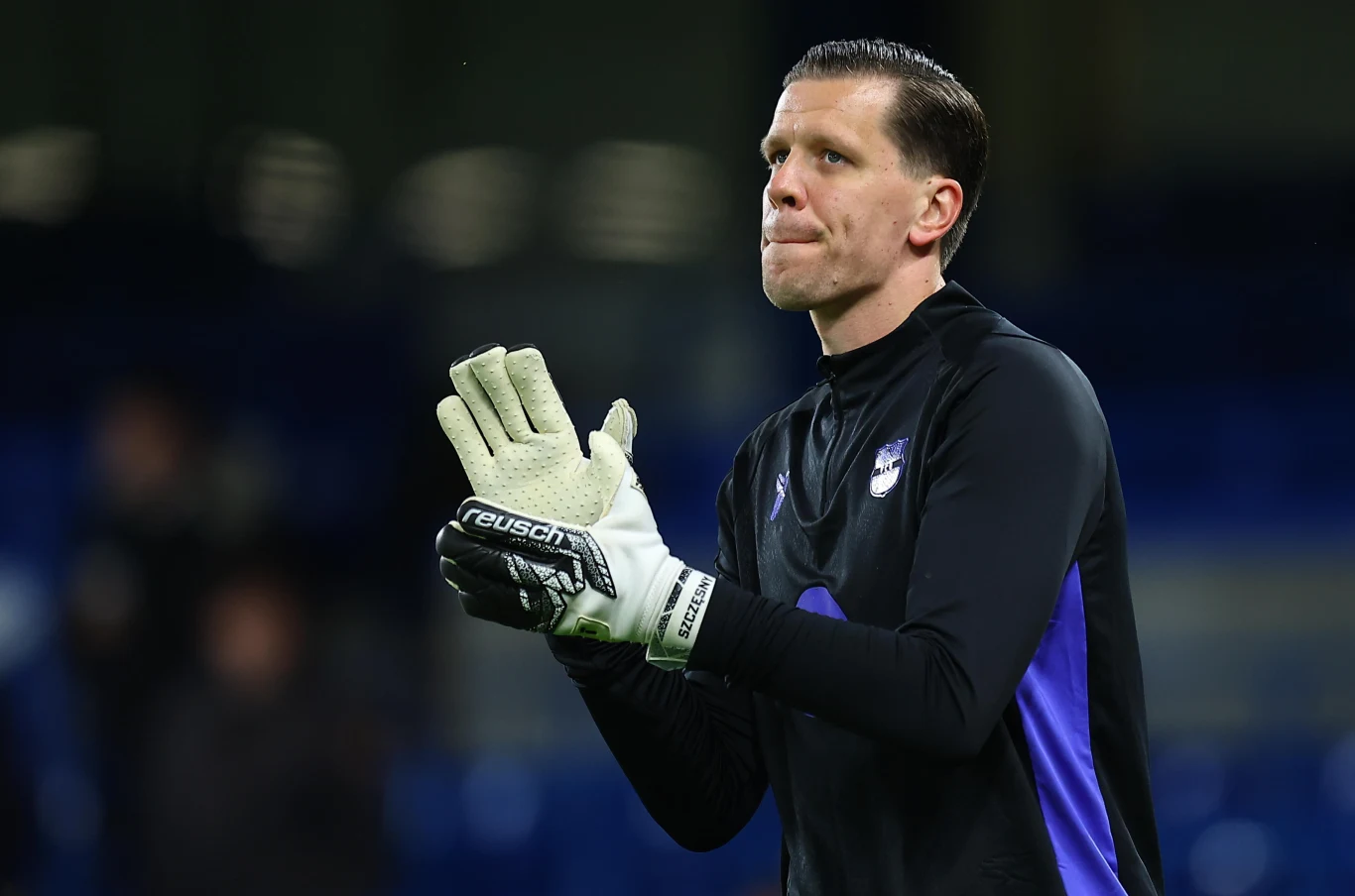 Piłkarz w czarnym stroju sportowym oraz rękawicach bramkarskich stoi na boisku i składa dłonie, patrząc przed siebie. W tle rozmazane niebieskie elementy stadionu.