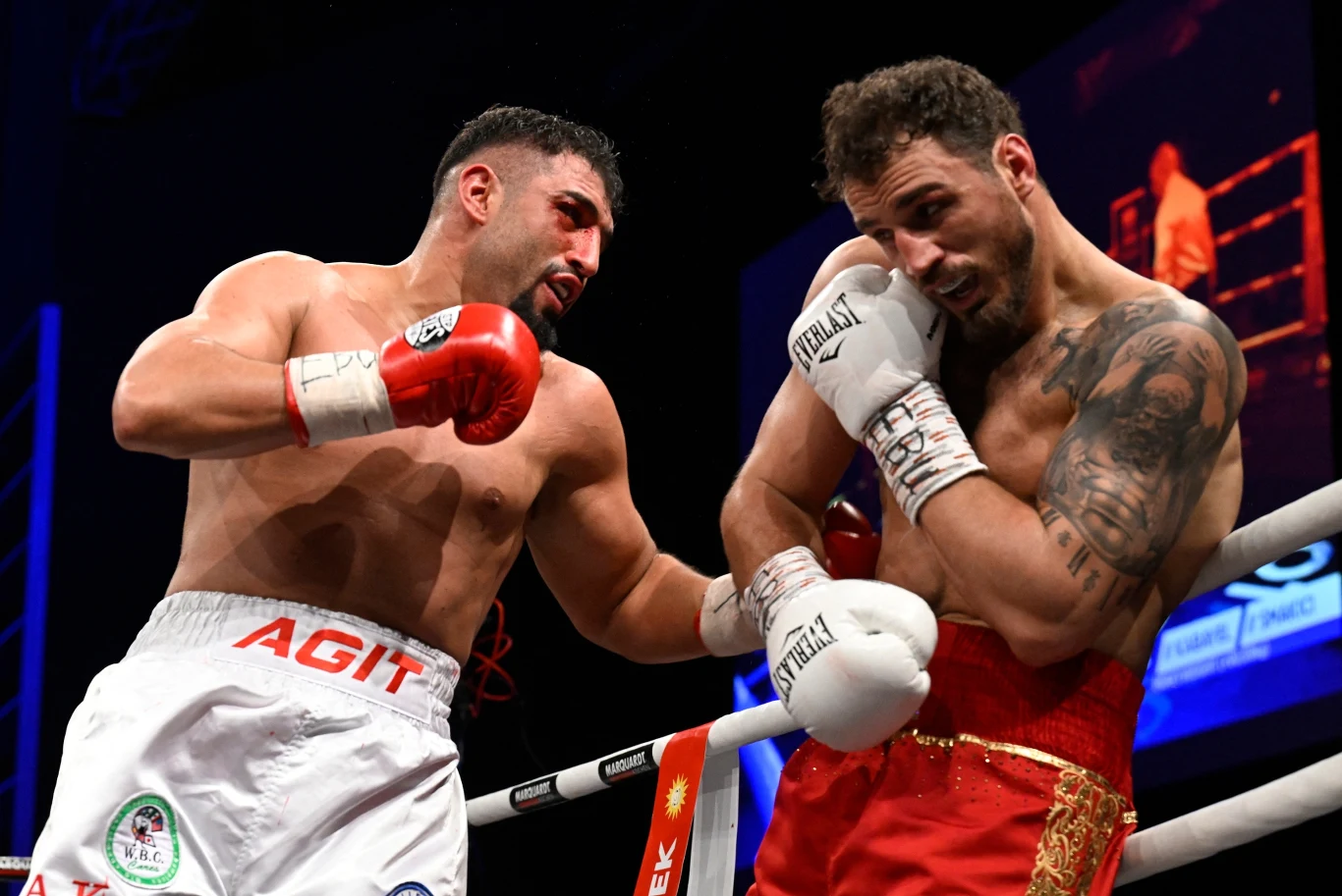 Dwóch bokserów podczas intensywnej walki na ringu, jeden z nich zadaje cios przeciwnikowi, który chroni się przy linach. Obaj zawodnicy w sportowych szortach, widoczne skupienie i wysiłek. Jasne światło podkreśla sylwetki bokserów, tłum i ring rozmazan...