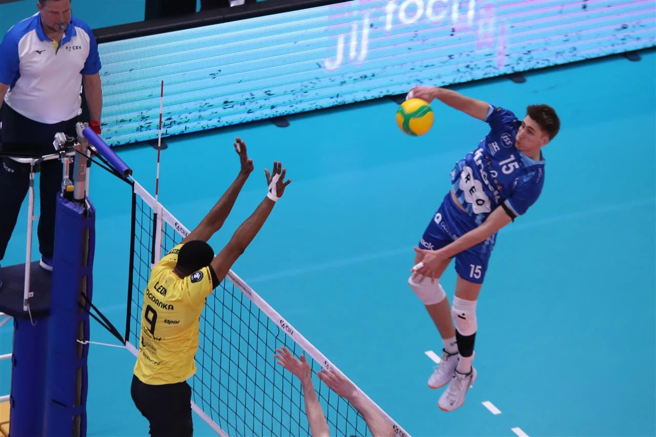 Wilfredo Leon w bloku w trakcie meczu z Knack Roeselare Siatkarz w niebieskim stroju wykonuje atak nad siatką, podczas gdy zawodnik w żółtej koszulce próbuje zablokować piłkę, obok nich stoi sędzia obserwujący przebieg akcji.