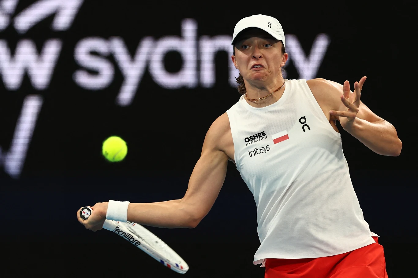 Tenisistka w dynamicznej pozie podczas wykonywania forhendowego uderzenia na korcie, skupiona na piłce, ubrana w biały sportowy top, czerwoną spódniczkę i białą czapkę, w tle rozmyte napisy i ciemne tło hali.