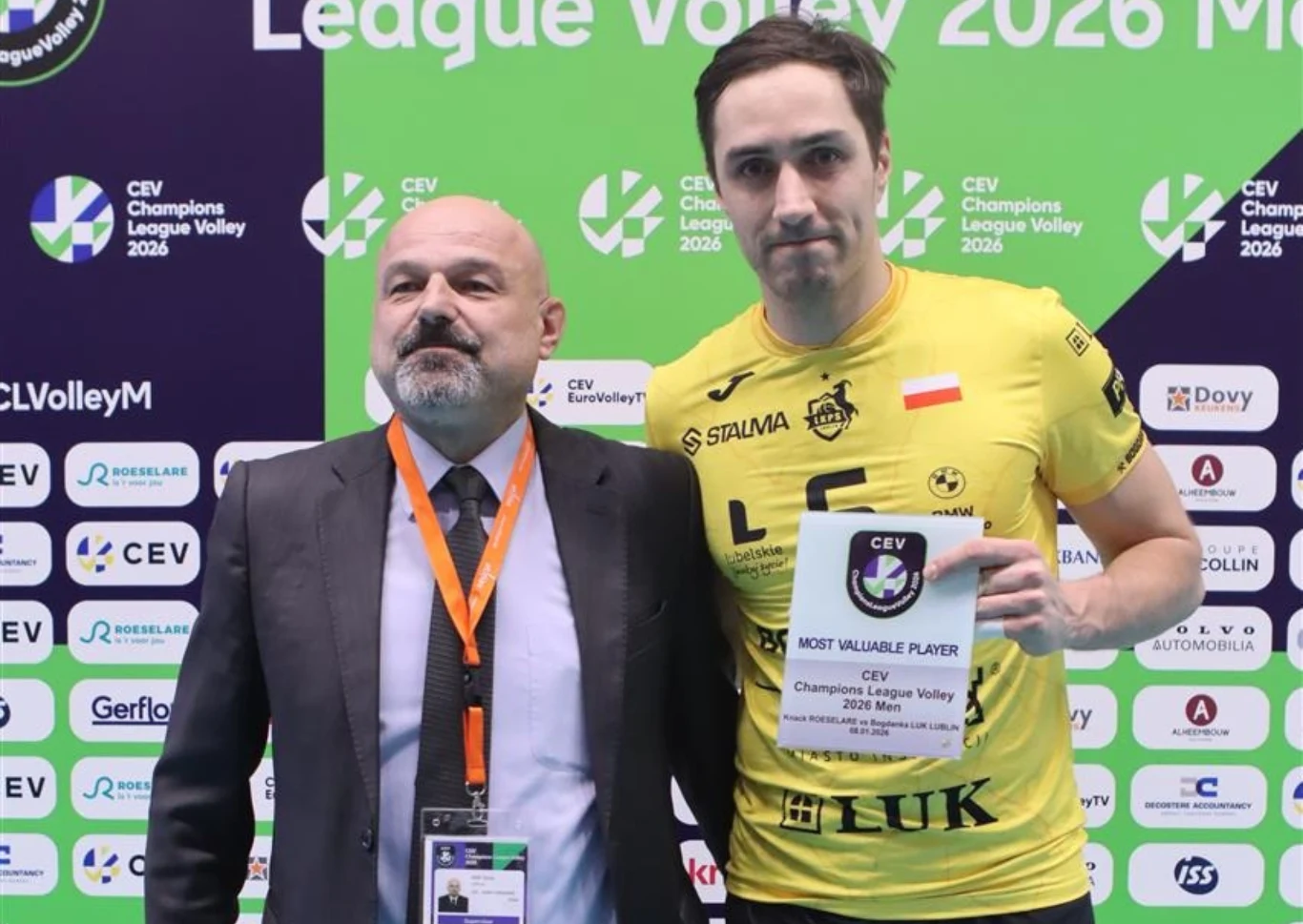 Mateusz Malinowski i jego "nagroda" za tytuł najlepszego zawodnika meczu Dwóch mężczyzn stojących razem na tle ścianki sponsorsko-turniejowej podczas wydarzenia siatkarskiego, jeden z nich ubrany w żółtą koszulkę sportową trzyma tabliczkę z napisem Most Valuable Player, drugi ma na sobie garnitur oraz zawieszkę z identyfika...