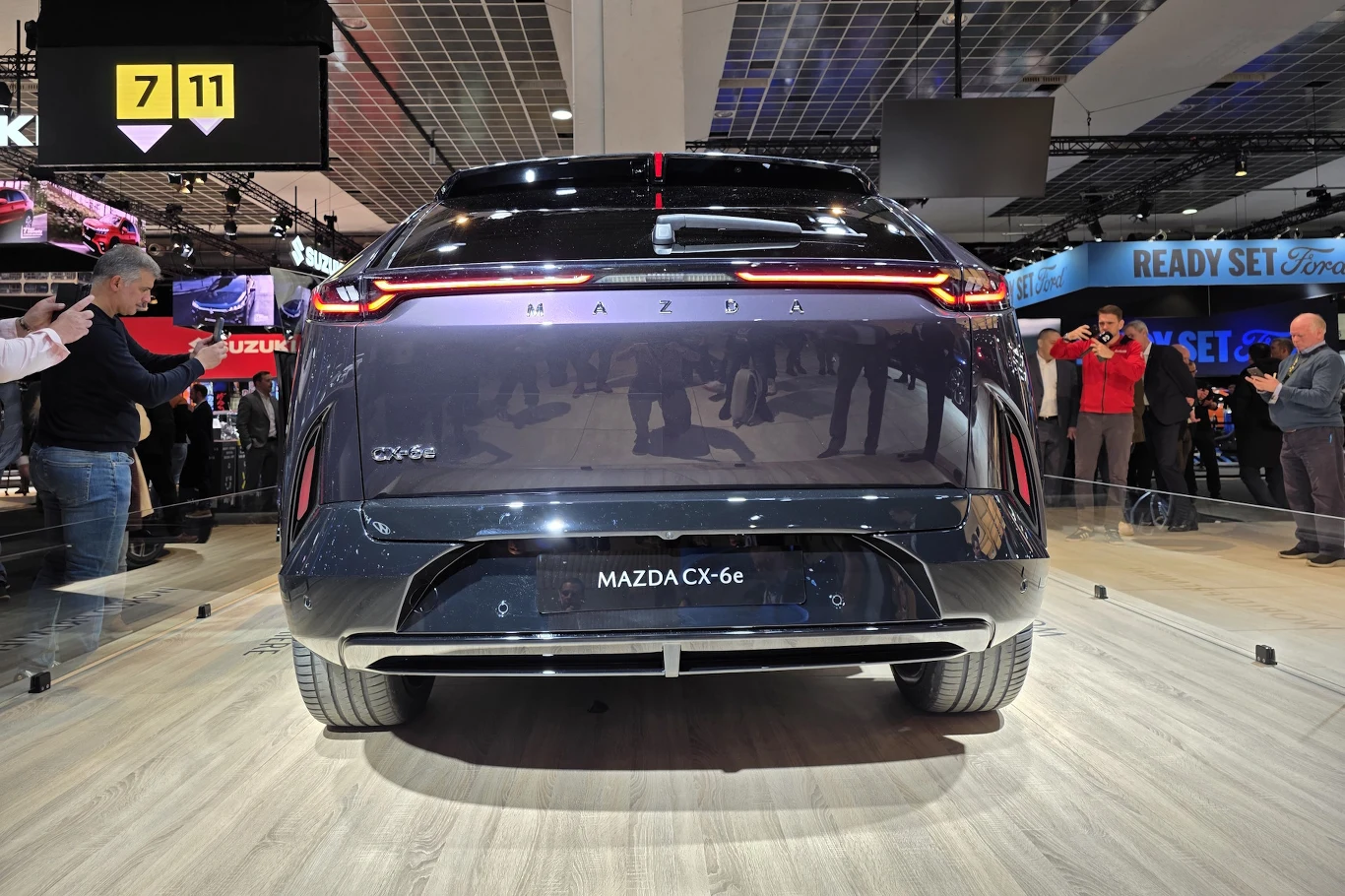 Mazda CX-6e Tylna część nowoczesnego samochodu Mazda CX-6e w ciemnym lakierze, ustawionego na ekspozycji targowej, wokół którego zgromadzeni są ludzie oraz fotografowie podczas prezentacji modelu.