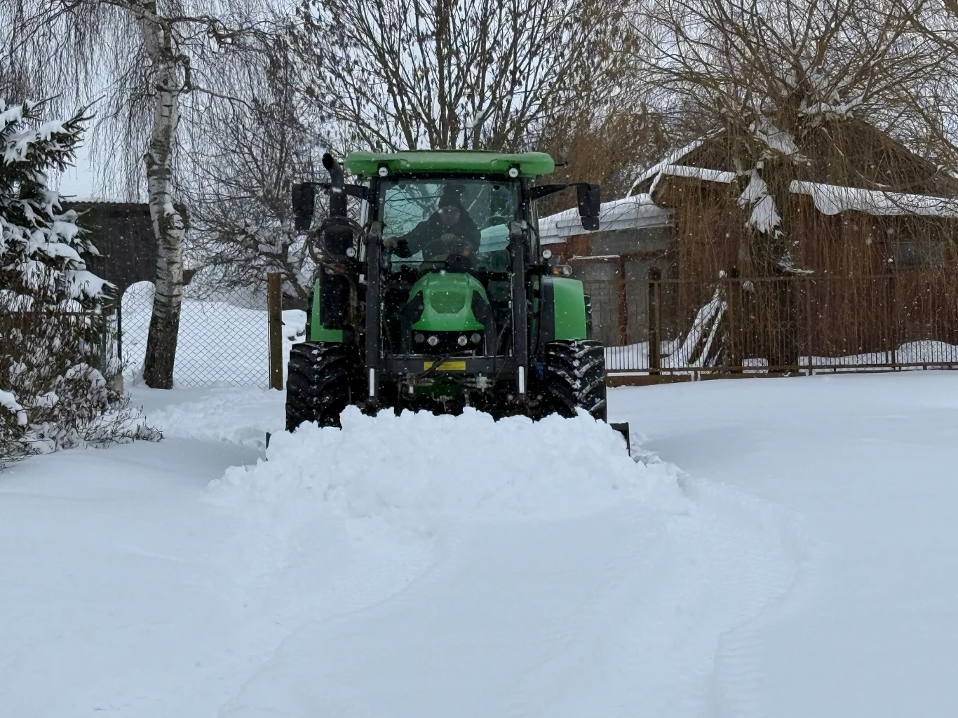 Zielony traktor usuwa śnieg z drogi na posesji, wokół leżą wysokie zaspy śniegu. W tle widoczne są zabudowania gospodarcze i drzewa pokryte śniegiem.
