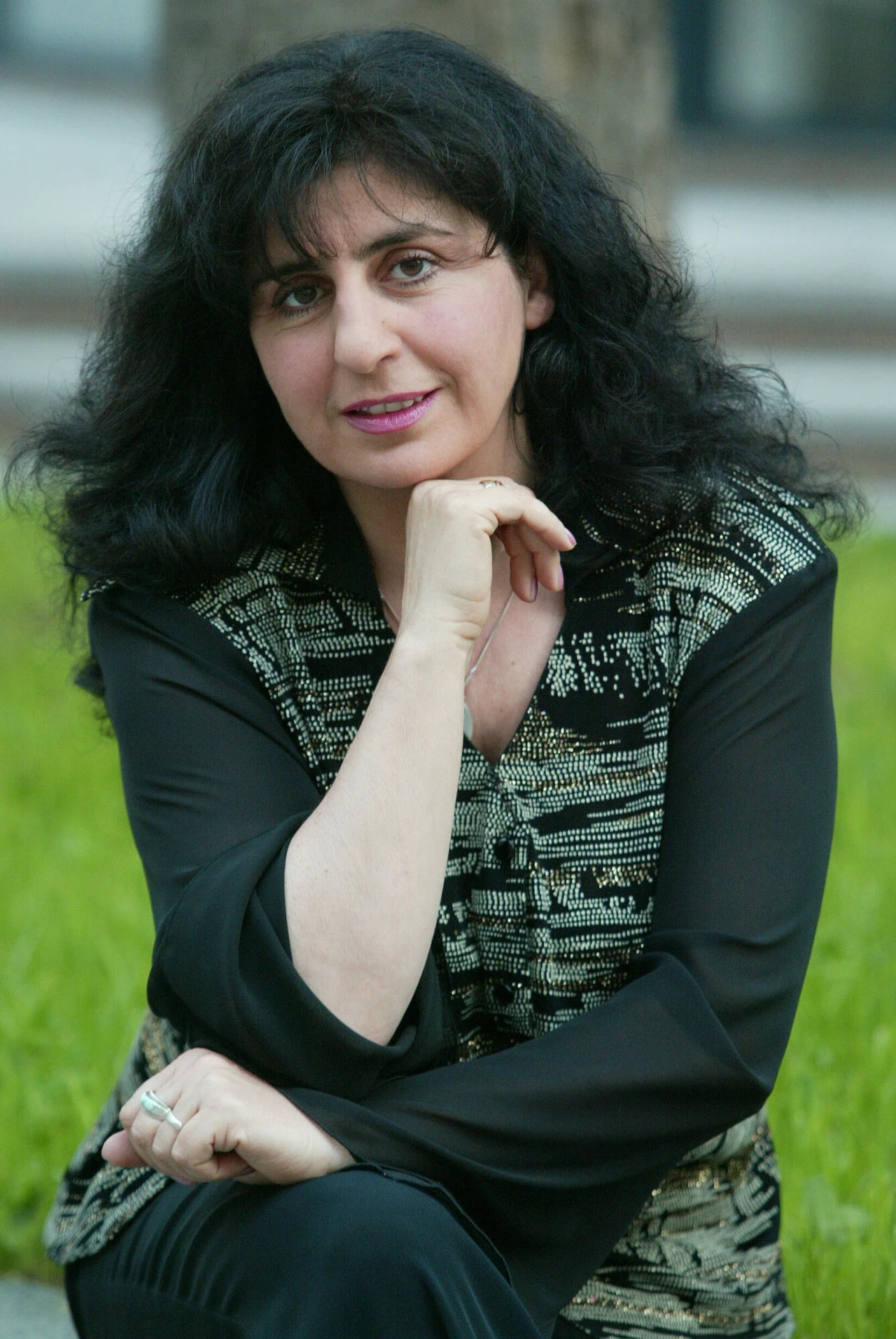 Kobieta z ciemnymi, kręconymi włosami siedzi na zewnątrz na tle zielonej trawy, opierając rękę na brodzie i uśmiechając się delikatnie do kamery.