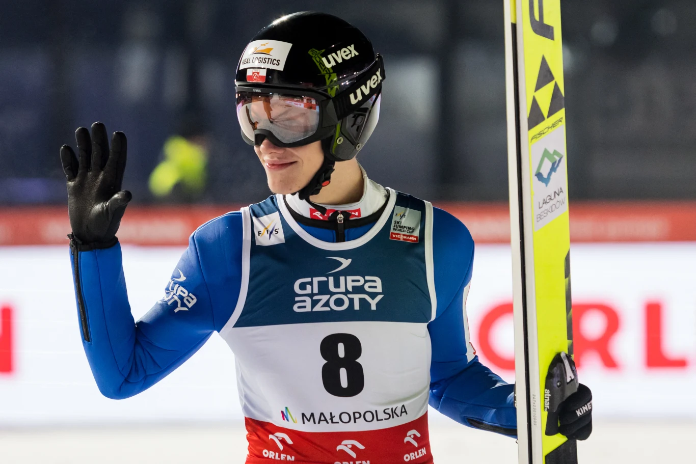 Skoczek narciarski ubrany w strój startowy z numerem 8, w kasku oraz goglach, trzyma narty i unosi dłoń w geście pozdrowienia, na tle reklamy sponsorów i areny sportowej.