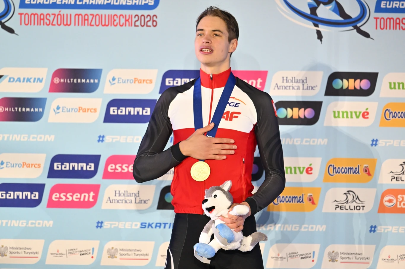 Młody sportowiec w stroju reprezentacyjnym z medalem na szyi, stojący z ręką na piersi, trzymający maskotkę, na tle ściany z logotypami sponsorów i napisem dotyczącym mistrzostw Europy w Tomaszowie Mazowieckim 2026.