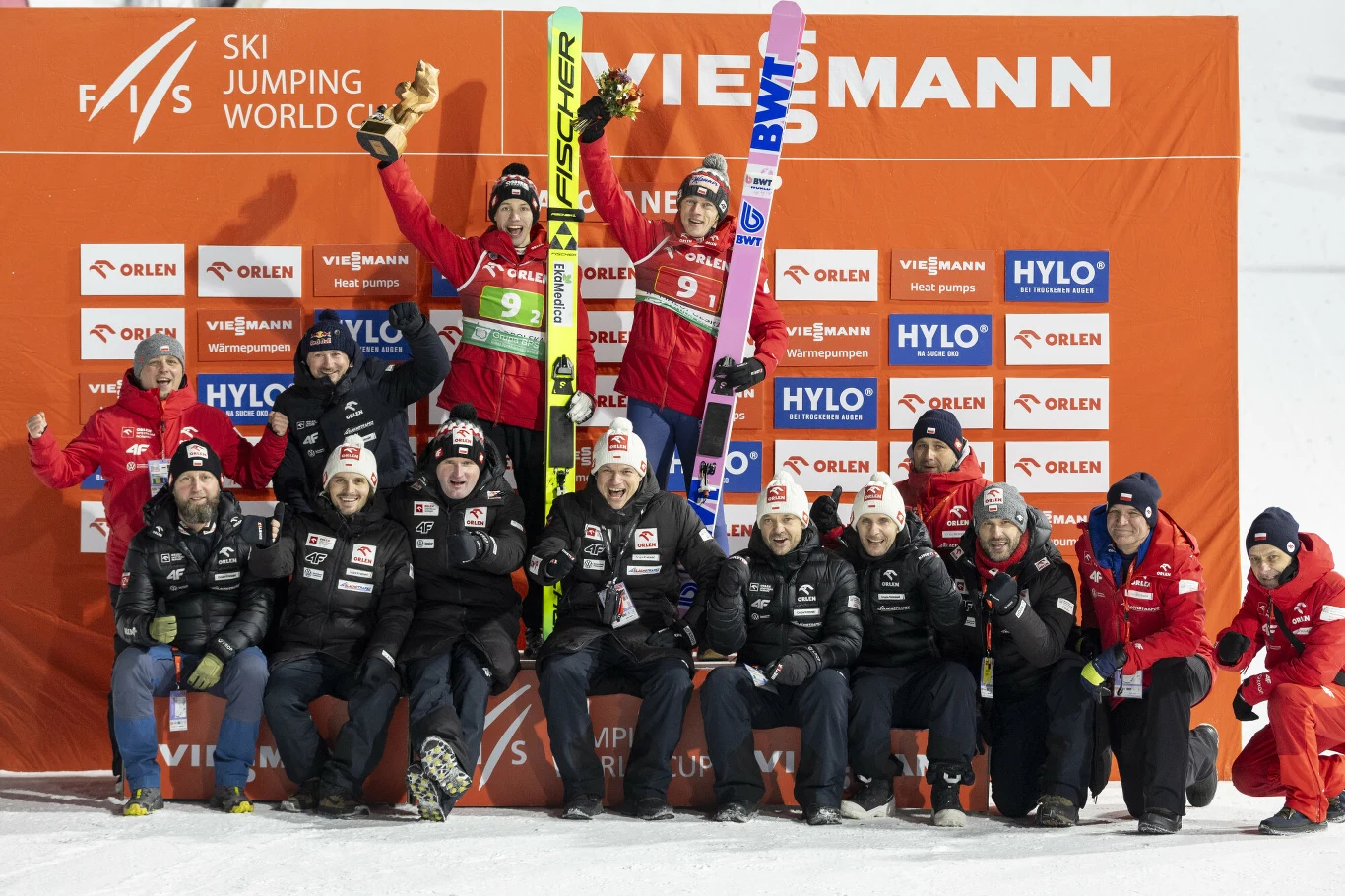 Grupa skoczków narciarskich wraz z zespołem technicznym pozuje na podium na tle pomarańczowej ścianki sponsorskiej, dwóch zawodników stoi z nartami uniesionymi w geście radości, pozostali członkowie zespołu siedzą i klęczą uśmiechając się do kamery.