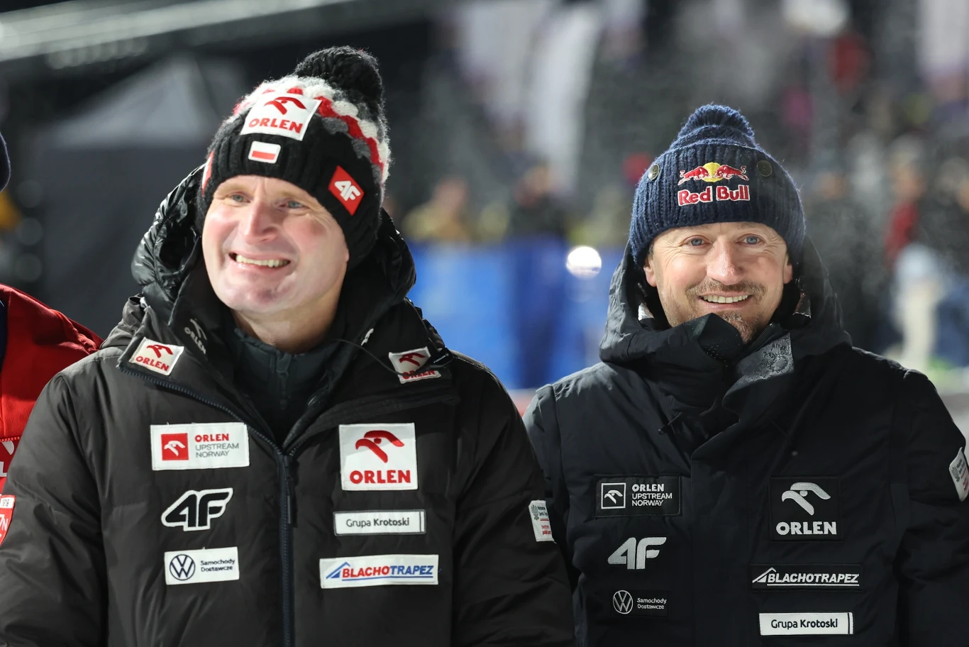 Dwóch mężczyzn ubranych w zimowe kurtki z logotypami sponsorów, stojących obok siebie i uśmiechających się, w tle rozmyte światła oraz nieokreślone osoby, zimowa atmosfera.