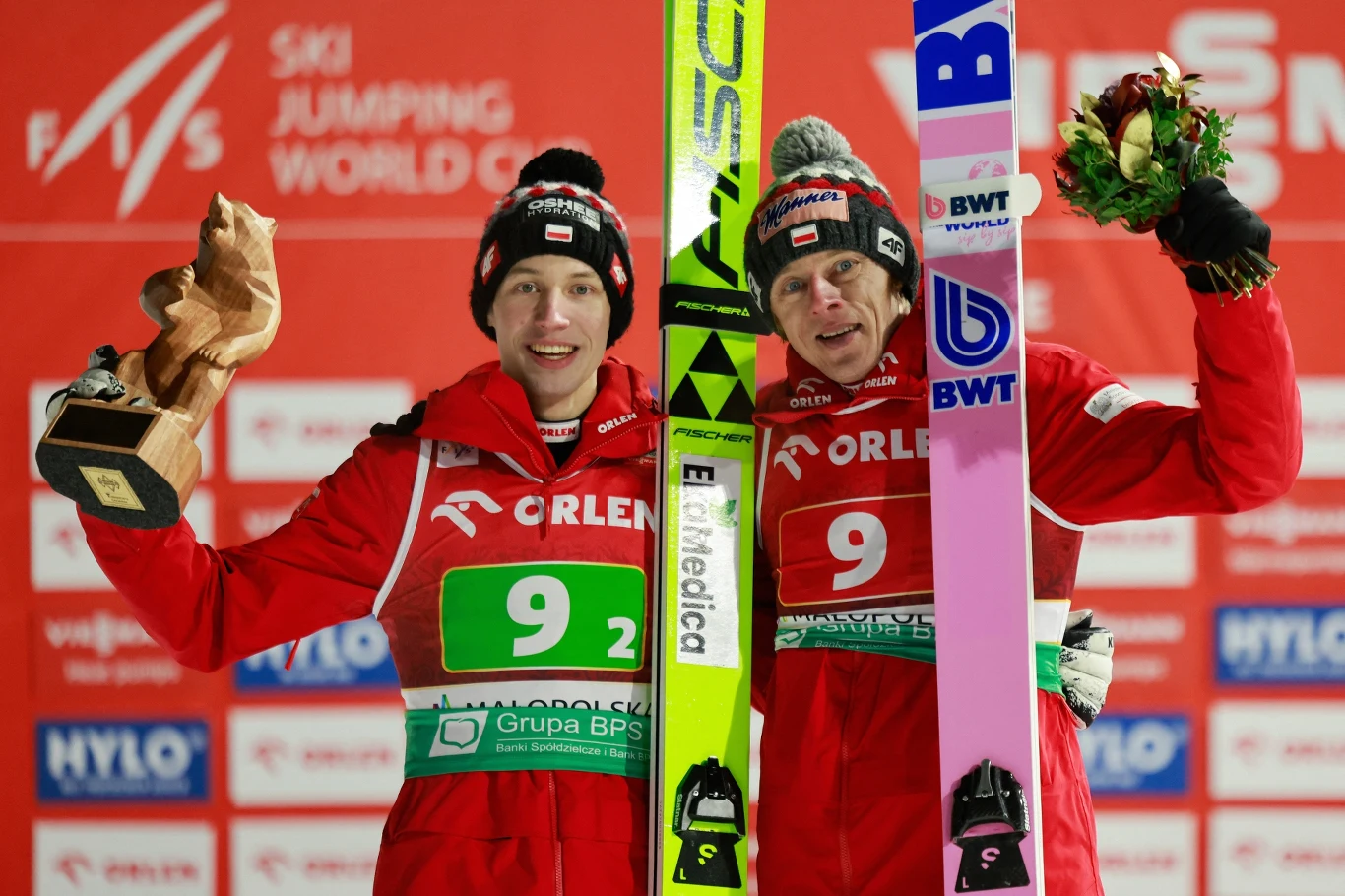 Dwóch polskich skoczków narciarskich stoi na podium z nartami, ubrani w czerwone kombinezony z logo sponsorów, jeden z nich trzyma drewnianą statuetkę, drugi bukiet kwiatów, w tle ścianka sponsora i logo zawodów Pucharu Świata.