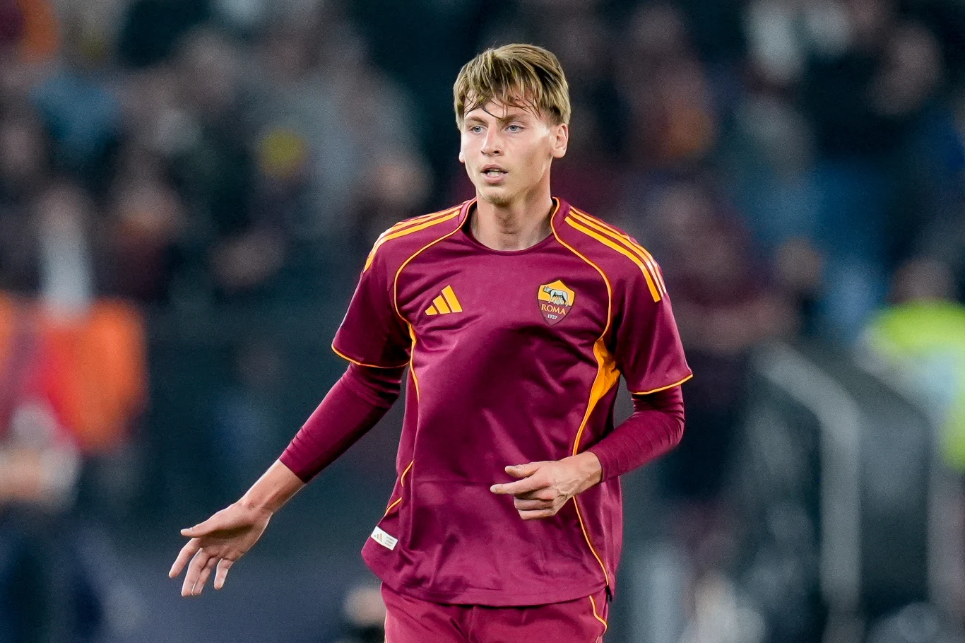 Piłkarz w bordowo-żółtym stroju drużyny AS Roma podczas meczu, skoncentrowany w trakcie gry na tle rozmytego stadionu i widzów.