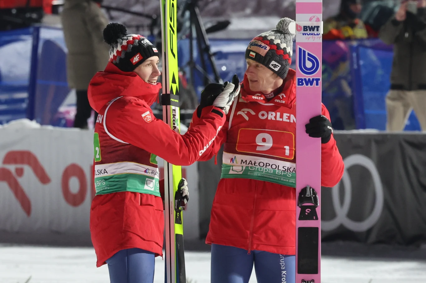 Dwóch skoczków narciarskich ubranych w czerwone kombinezony i czapki zimowe rozmawia ze sobą na skoczni. Jeden z nich trzyma narty, w tle widać elementy infrastruktury sportowej oraz innych uczestników zawodów.