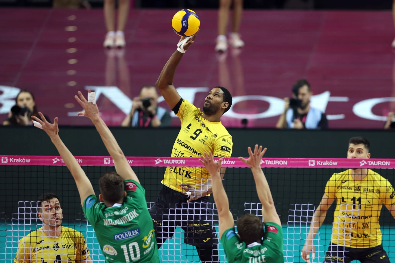 Zawodnik drużyny w żółtych koszulkach wykonuje atak nad siatką podczas meczu siatkówki, podczas gdy dwóch graczy w zielonych koszulkach próbuje go zablokować. W tle widać innych zawodników oraz fragment hali sportowej.