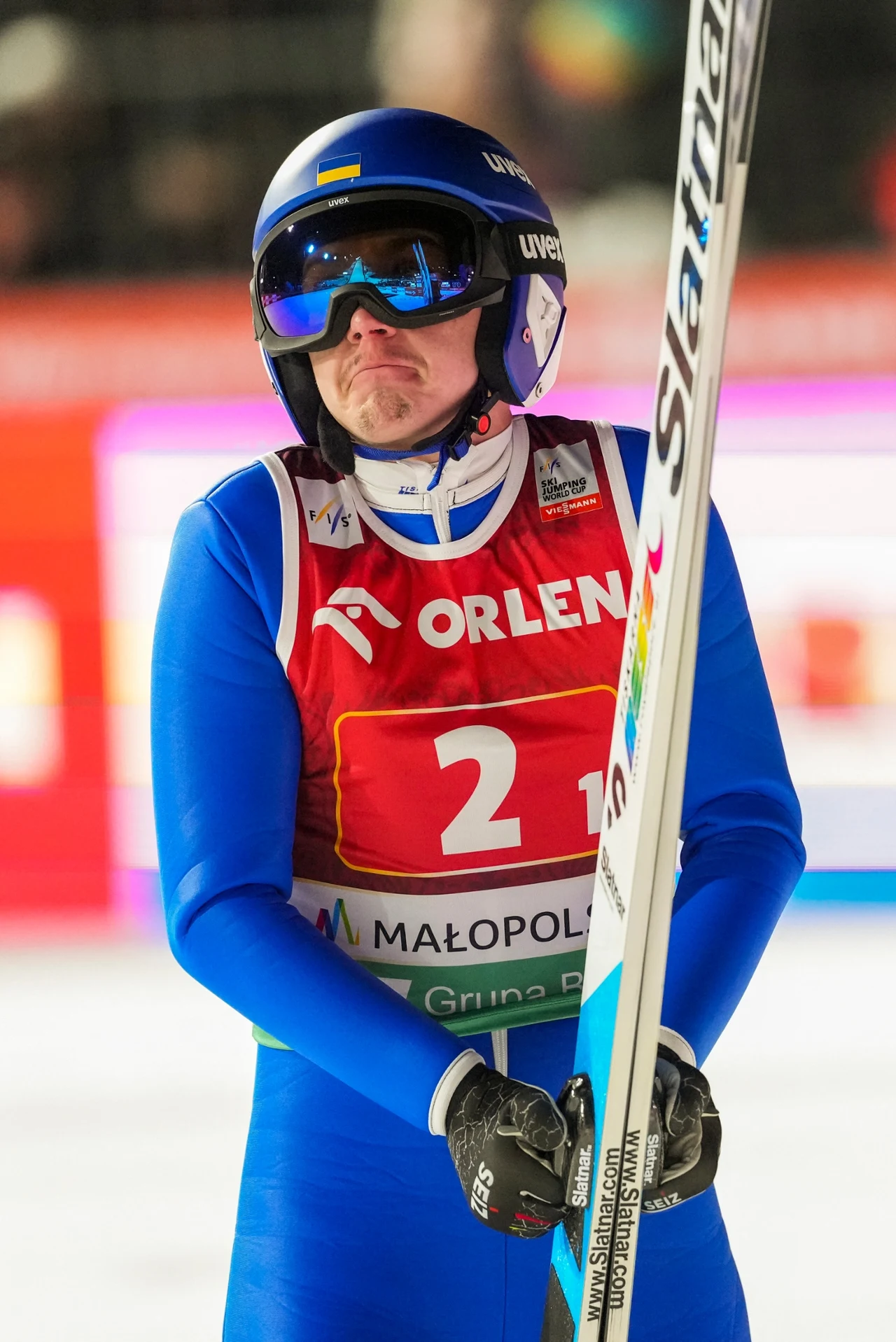 Zawodnik w kasku i goglach narciarskich w niebieskim kombinezonie, trzymający narty podczas zawodów skoków narciarskich, wyraz twarzy sugeruje rozczarowanie lub niezadowolenie z oddanego skoku.