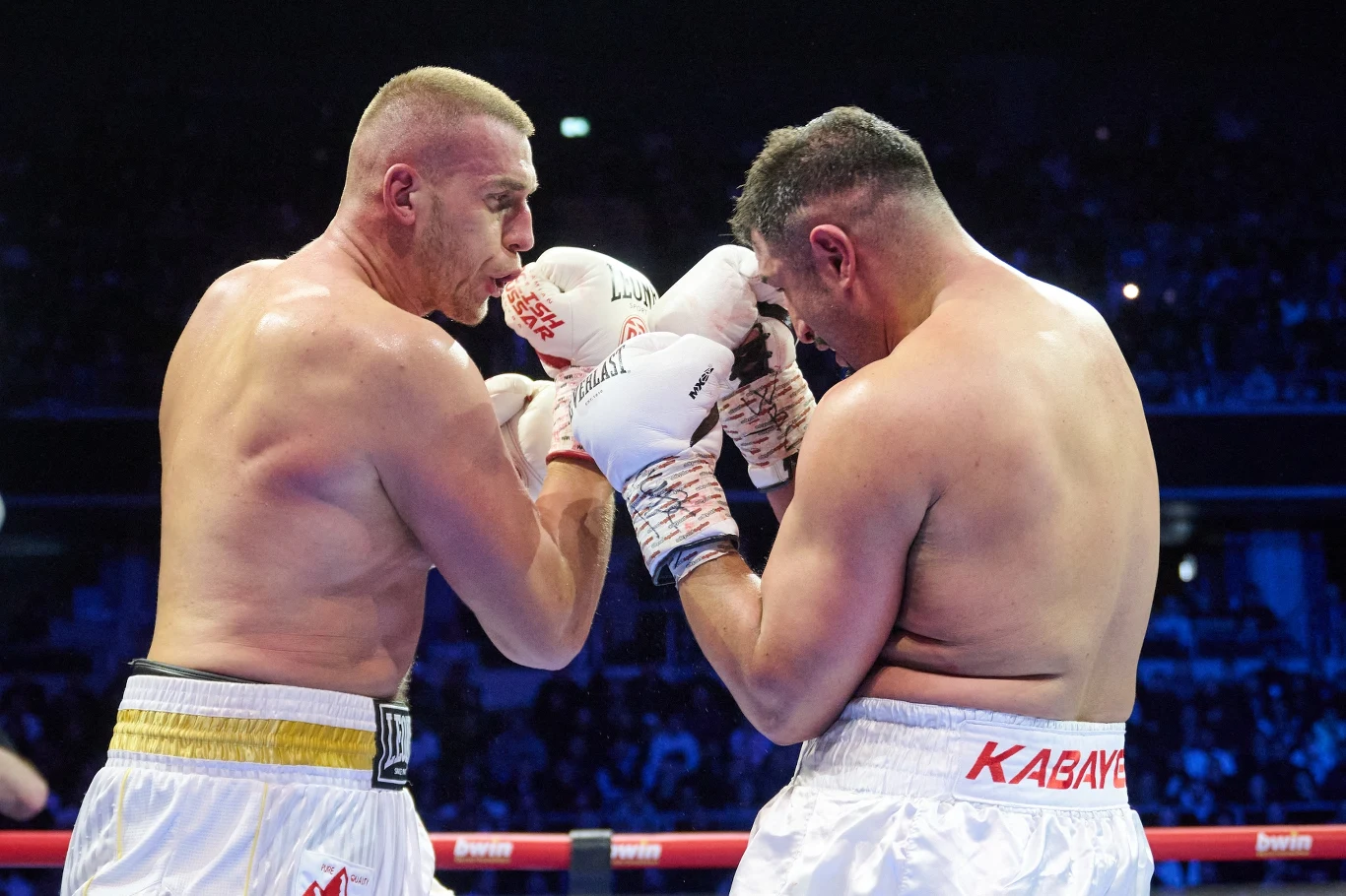 Dwóch bokserów w ringu podczas intensywnej wymiany ciosów, obaj skupieni, z napiętymi mięśniami, w sportowych szortach i rękawicach, wokół publiczność oświetlona światłami areny.