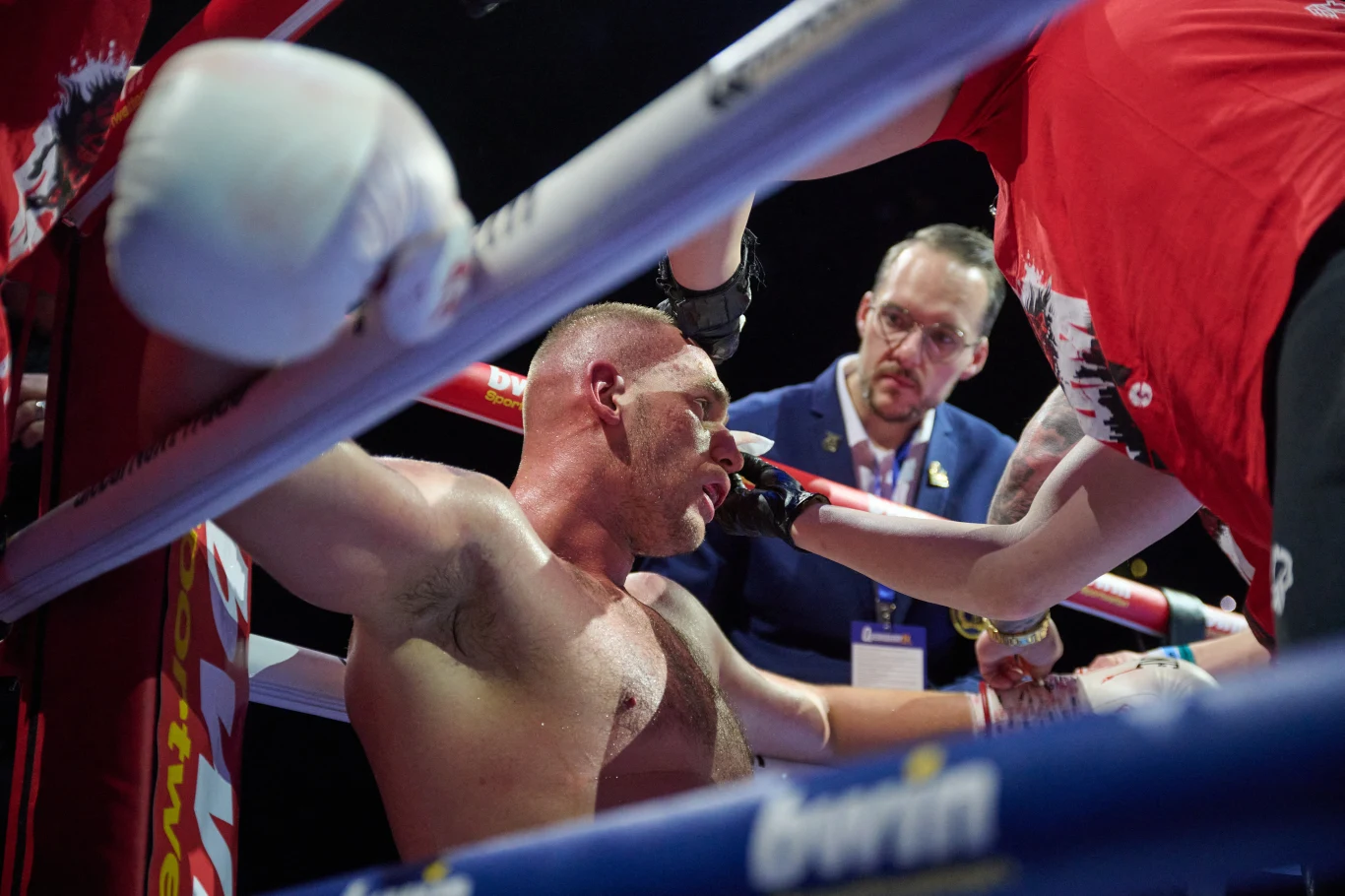 Bokser siedzący na ringu w trakcie przerwy, otoczony przez członków swojego sztabu, którzy udzielają mu wskazówek i wsparcia, w tle sędzia obserwujący sytuację.