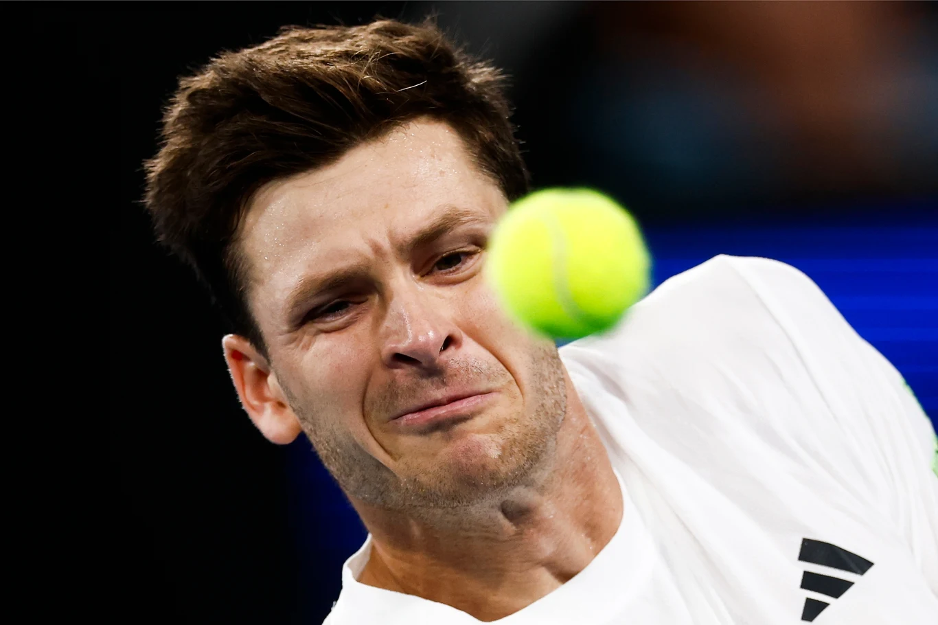 Zbliżenie twarzy tenisisty podczas intensywnej gry z lecącą żółtą piłką tenisową tuż przed jego oczami, wyraz skupienia i determinacji widoczny na twarzy sportowca, ubranie sportowe z widocznym logo.