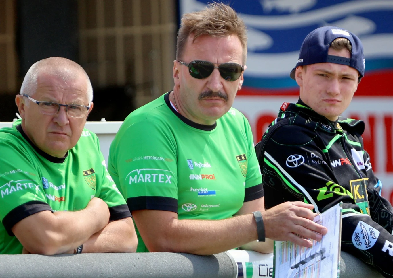 Trzech mężczyzn ubranych w zielone sportowe koszulki i ciemny sportowy kombinezon stoi obok siebie, patrząc w jednym kierunku, jeden z nich trzyma dokumenty lub notatki.