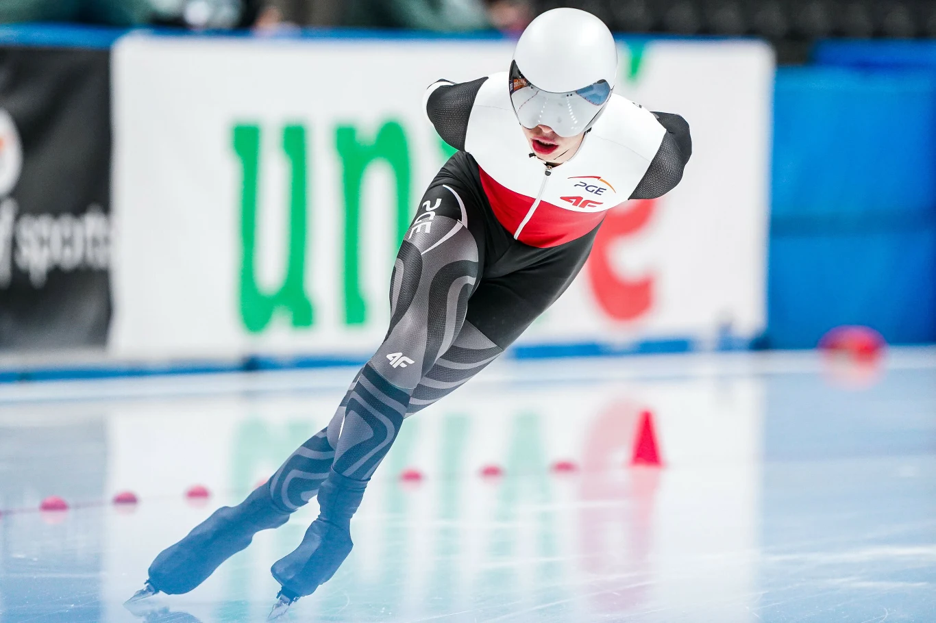 Łyżwiarz szybki w kombinezonie startowym w dynamicznym pochyleniu podczas jazdy po lodowisku, wokół rozmieszczone czerwone znaczniki trasy, w tle rozmyte trybuny i banery reklamowe.