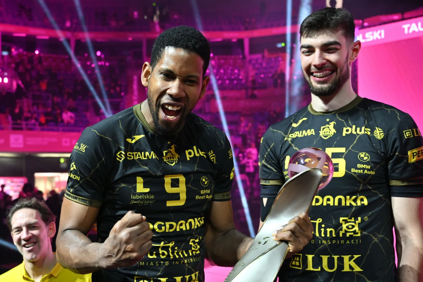 Dwóch siatkarzy ubranych w czarne koszulki z żółtymi napisami świętuje zwycięstwo na arenie sportowej, jeden z nich trzyma trofeum i zaciska pięść w geście radości, w tle widoczne światła i fragment trybun.