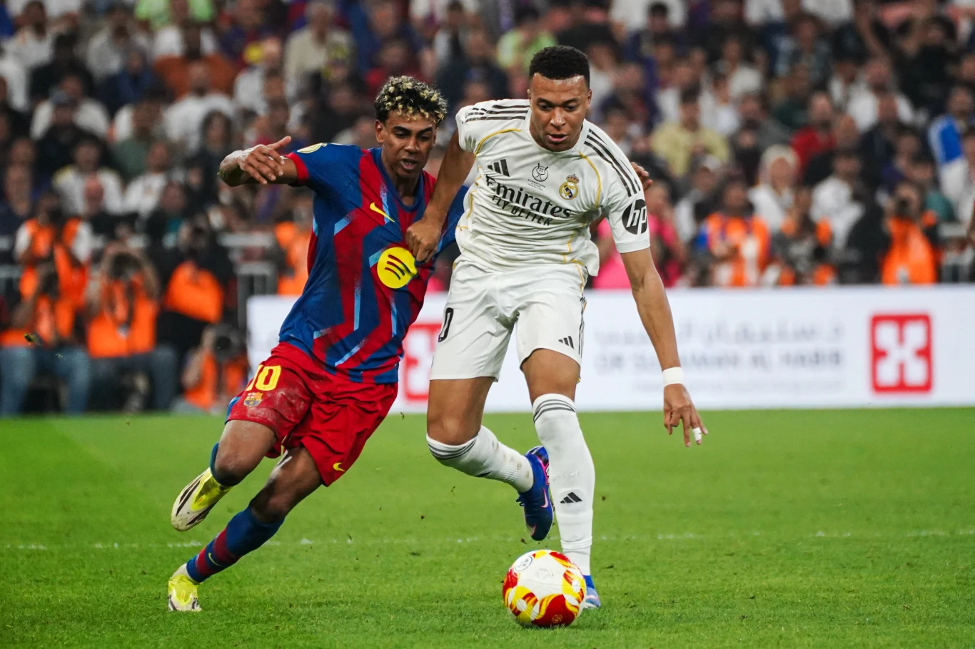 Dwóch piłkarzy walczy o piłkę na murawie podczas meczu, jeden z zawodników ubrany w białą koszulkę Realu Madryt, drugi w niebiesko-czerwonym stroju Barcelony, w tle widać kibiców oraz rozmyty stadion.