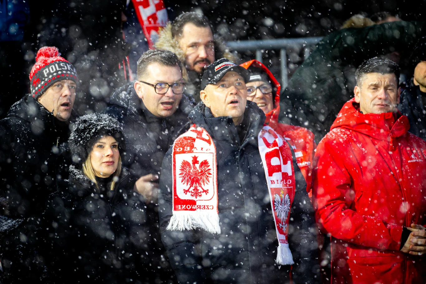 Grupa kibiców ubrana w czerwone i białe szaliki oraz kurtki, stojąca razem podczas padającego śniegu, okazująca wsparcie swojej drużynie narodowej.