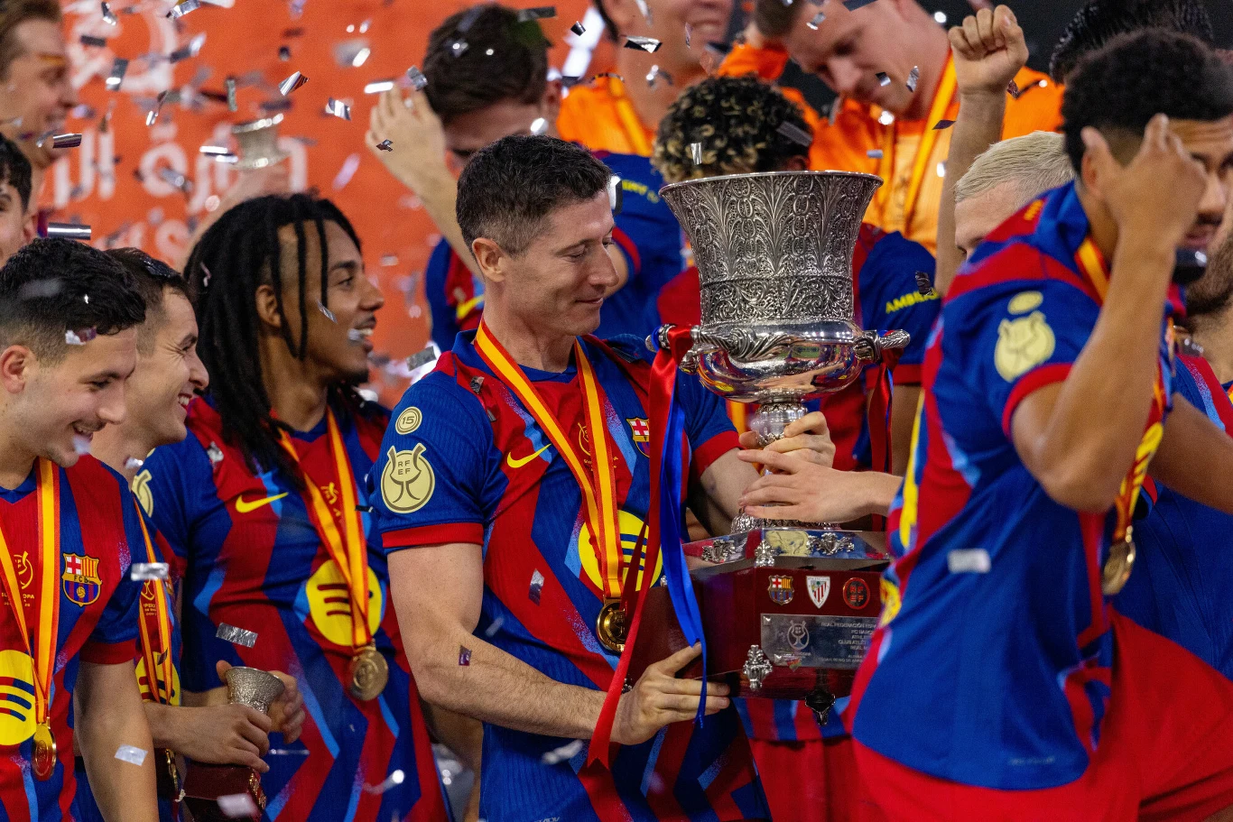 Piłkarze drużyny FC Barcelona w barwnych strojach świętują zwycięstwo, trzymając dużych rozmiarów puchar, wokół nich unoszą się konfetti, a na szyjach mają zawieszone złote medale.