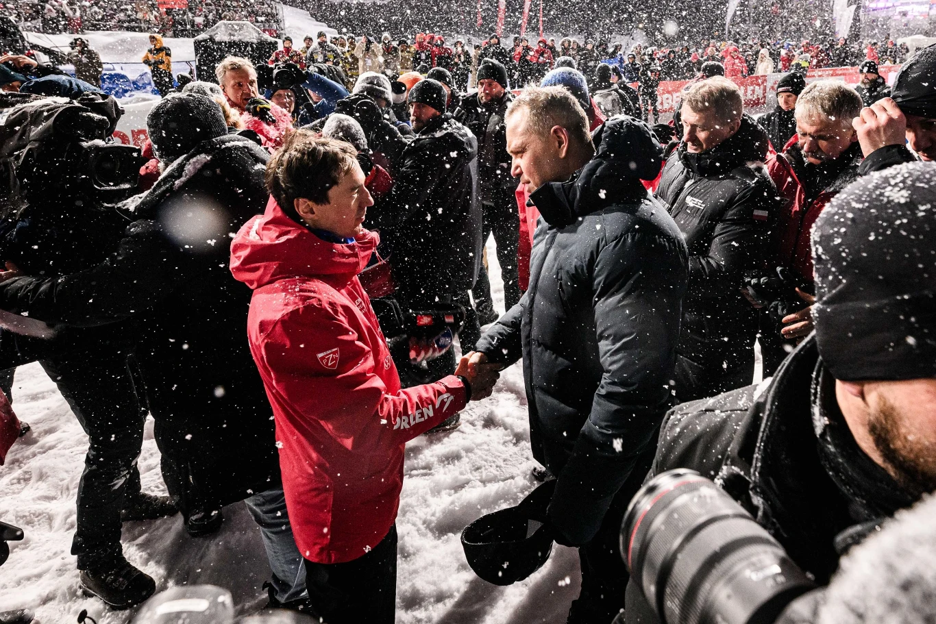 Kamil Stoch i Karol Nawrocki Tłum ludzi zgromadzony na zewnątrz podczas intensywnych opadów śniegu. Dwóch mężczyzn w centrum kadru uściskiem dłoni wymienia pozdrowienia; jeden ubrany w czerwoną kurtkę, drugi w czarną. W tle widoczne postacie w zimowych ubraniach oraz reporterska k...