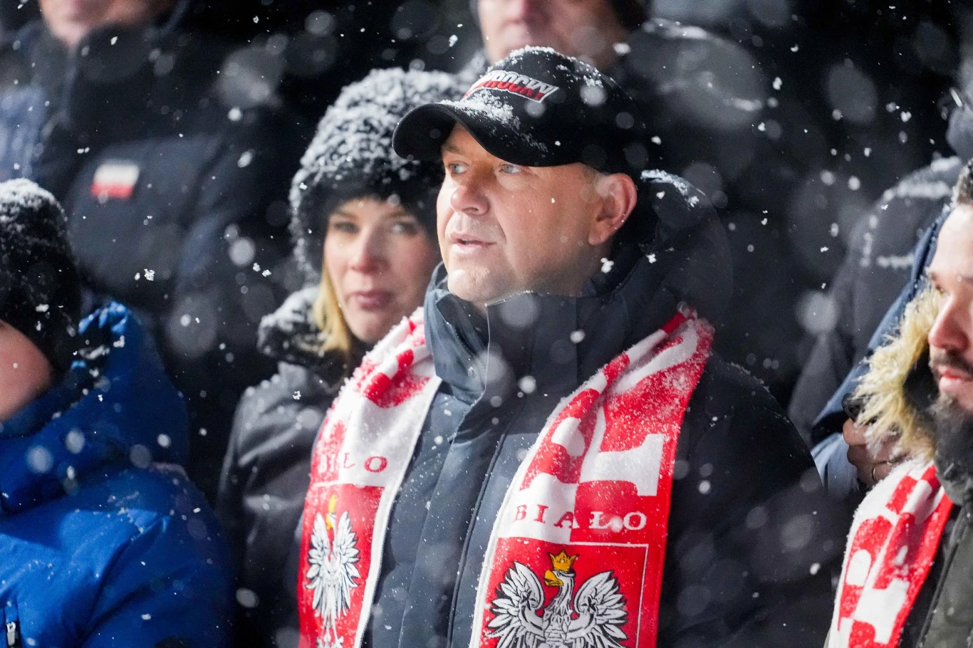 Prezydent na konkursie PŚ w Zakopanem Mężczyzna w czapce z daszkiem oraz biało-czerwonym szaliku z godłem Polski stoi w śniegu w tłumie ludzi ubraną w zimowe kurtki, wokół niego widoczna padająca śnieżyca.