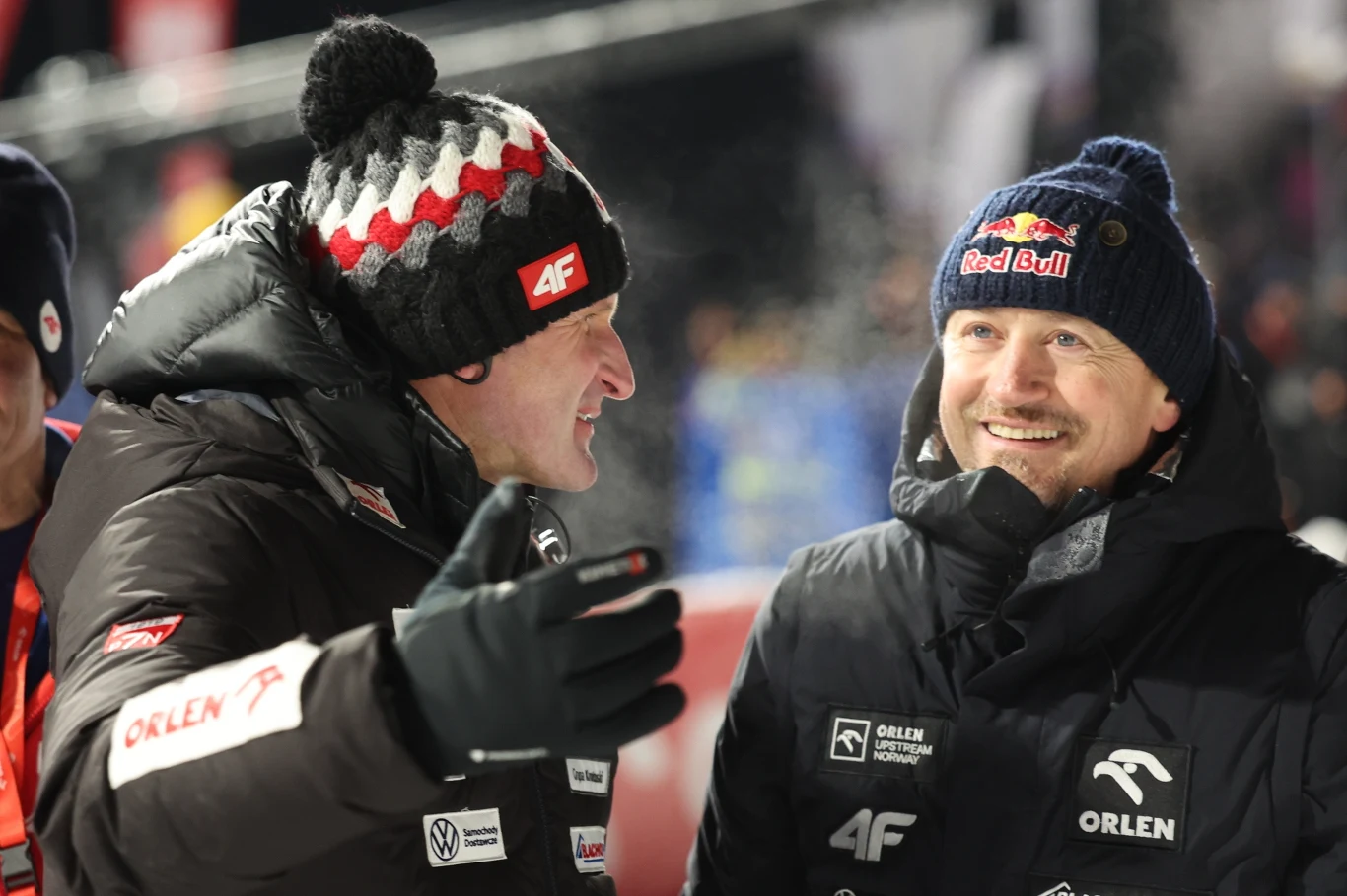 Maciej Maciusiak (po lewej) i Adam Małysz Dwóch mężczyzn uśmiechniętych rozmawia ze sobą, ubrani w zimowe kurtki z logo sponsorów oraz czapki, jeden w czapce Red Bull, drugi w charakterystycznym nakryciu głowy marki 4F, tło rozmyte sugerujące wydarzenie sportowe na zewnątrz.