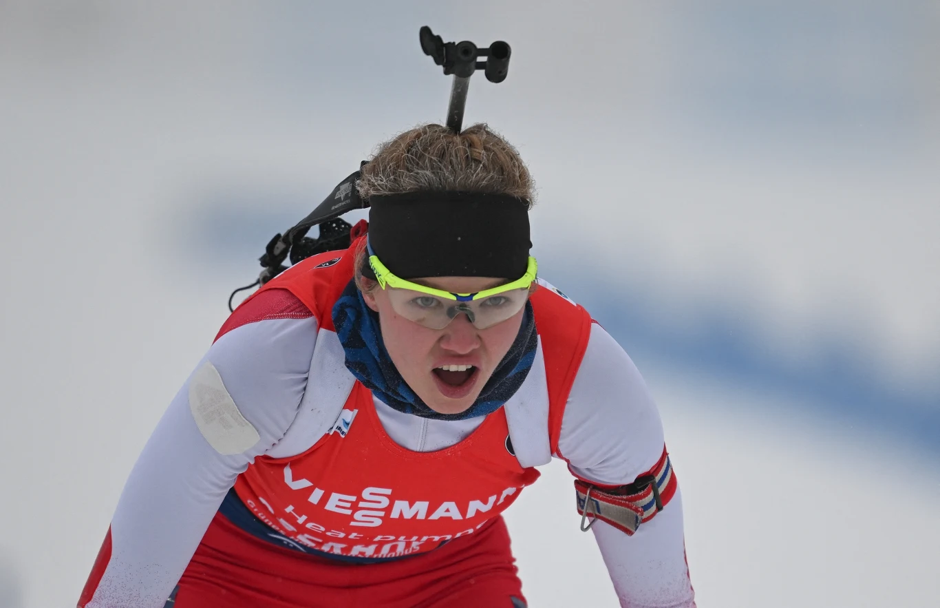 Daria Dalidowicz Zawodniczka w stroju sportowym biegnie na nartach w trakcie zawodów biathlonowych, na plecach niesie karabin, na twarzy wyraz determinacji, kontrastujące okulary sportowe dodają dynamiki scenie.