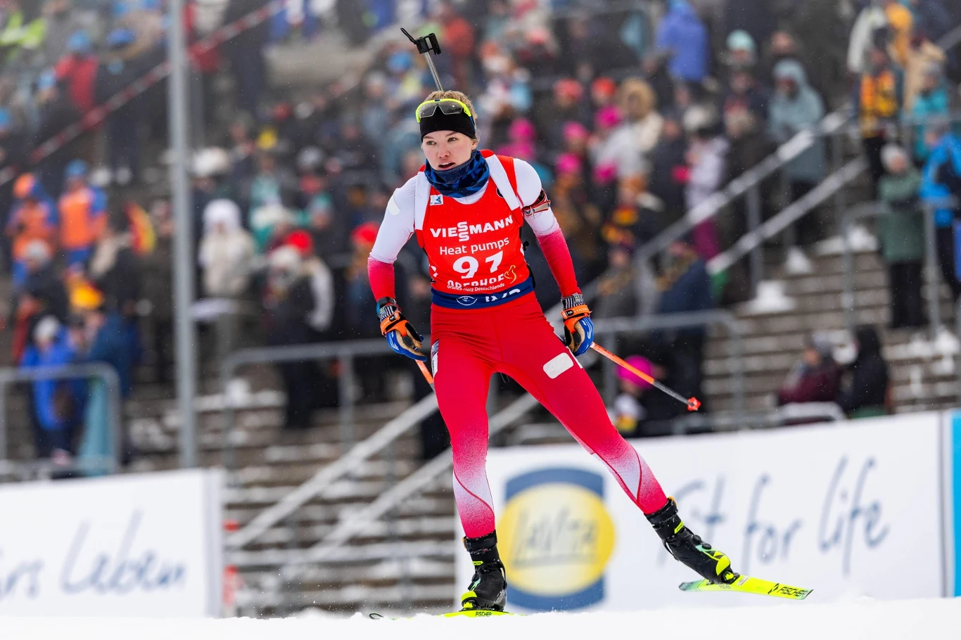 Daria Dalidowicz Zawodniczka biathlonowa w czerwono-białym stroju sportowym podczas zawodów, na plecach niesie karabin, wokół ośnieżone trybuny i kibice w tle.