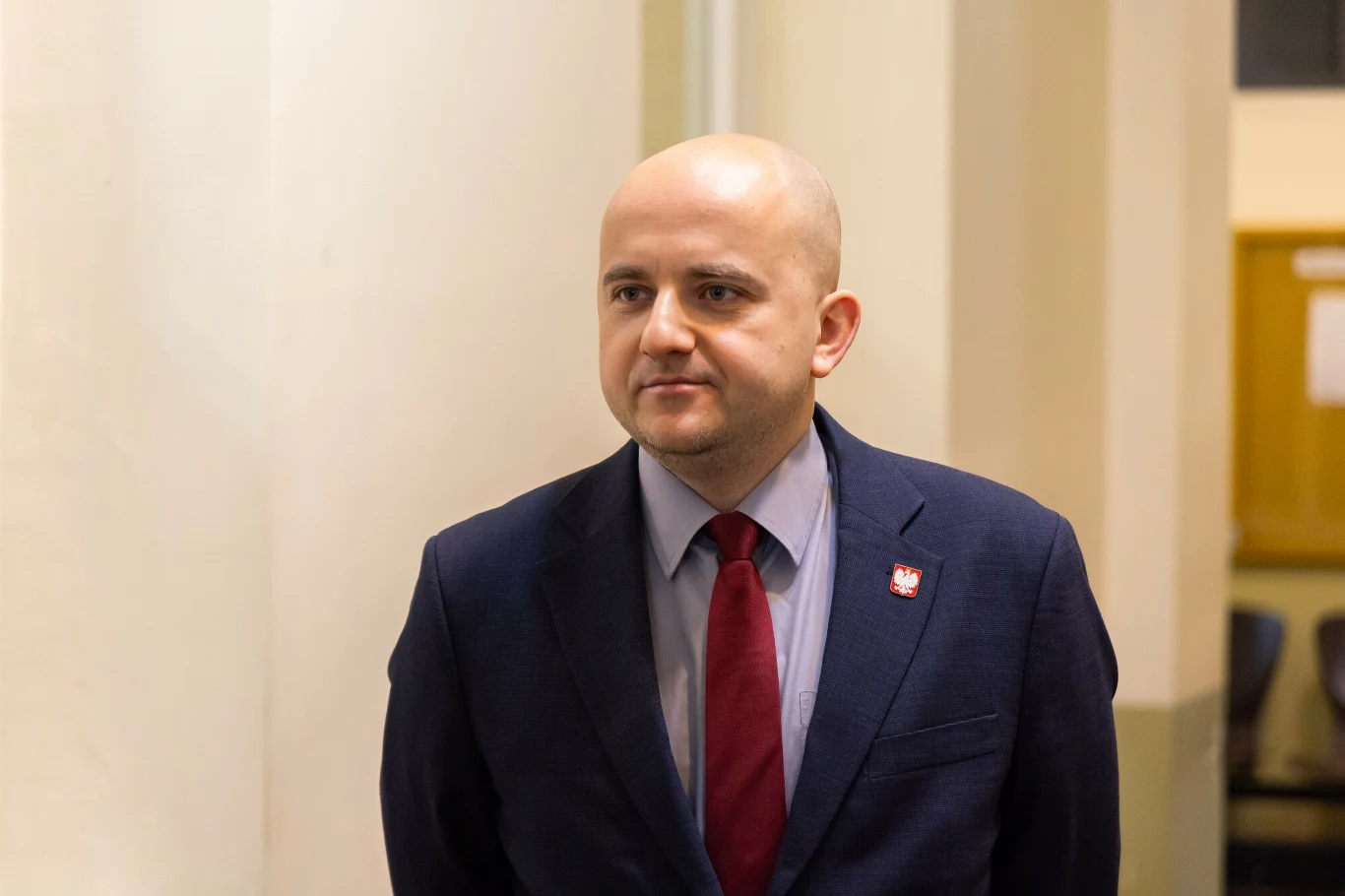 Mężczyzna w garniturze i czerwonym krawacie stoi w jasnym wnętrzu, przypięta do klapy marynarki odznaka z godłem Polski, w tle fragment kolumny i tablica informacyjna.