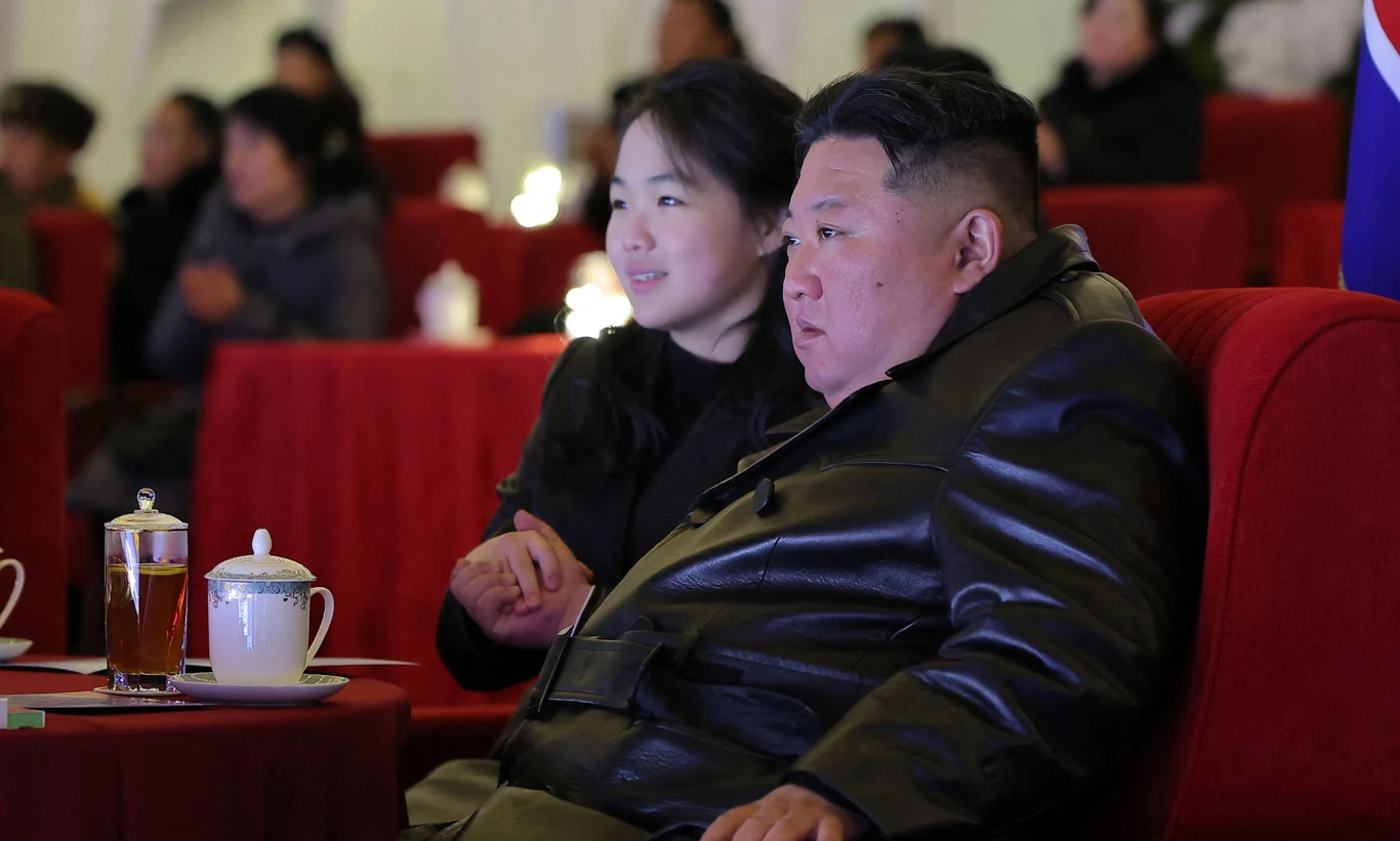 Kim Dzong Un wraz ze swoją córką Kim Ju Ae. Zdjęcie opublikowane w grudniu 2025 przez państwowe media Mężczyzna w czarnym skórzanym płaszczu siedzi obok kobiety na czerwonym fotelu w eleganckim otoczeniu, na stoliku obok stoi filiżanka z herbatą pod przykryciem i szklanka z napojem. W tle widoczni są inni uczestnicy wydarzenia.