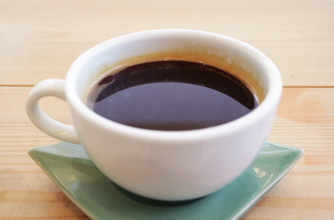 Biała filiżanka wypełniona czarną kawą ustawiona na jasnozielonym spodku, całość na jasnym drewnianym stole.