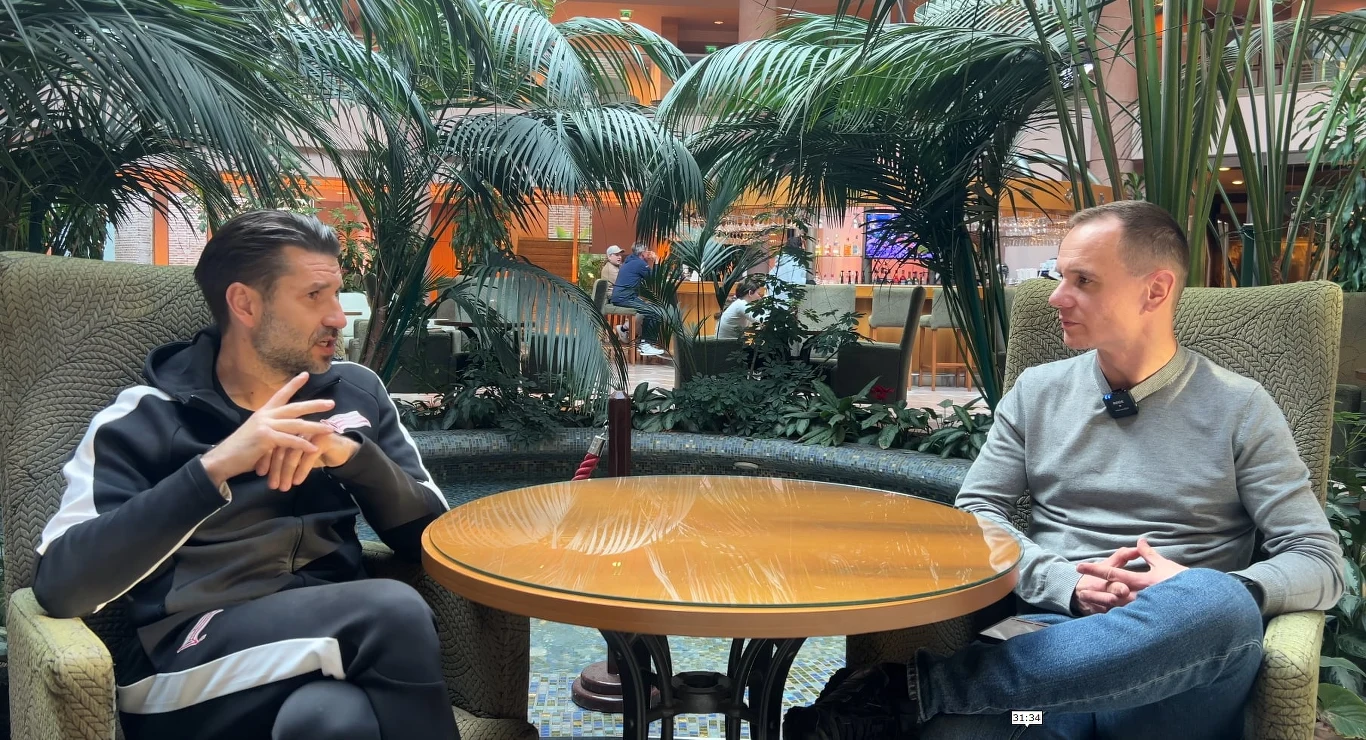 Dwóch mężczyzn siedzących naprzeciwko siebie przy okrągłym stole, rozmawiających w przestronnym wnętrzu z bujną roślinnością w tle. Obaj są zrelaksowani, a otoczenie sprawia wrażenie swobodnej atmosfery.