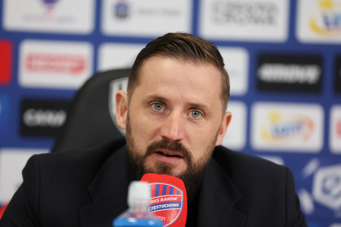 Mężczyzna z krótką brodą i zaczesanymi włosami udziela wywiadu podczas konferencji prasowej, siedząc przy biurku z mikrofonem, w tle widoczne są logotypy sponsorów i organizacji sportowej.