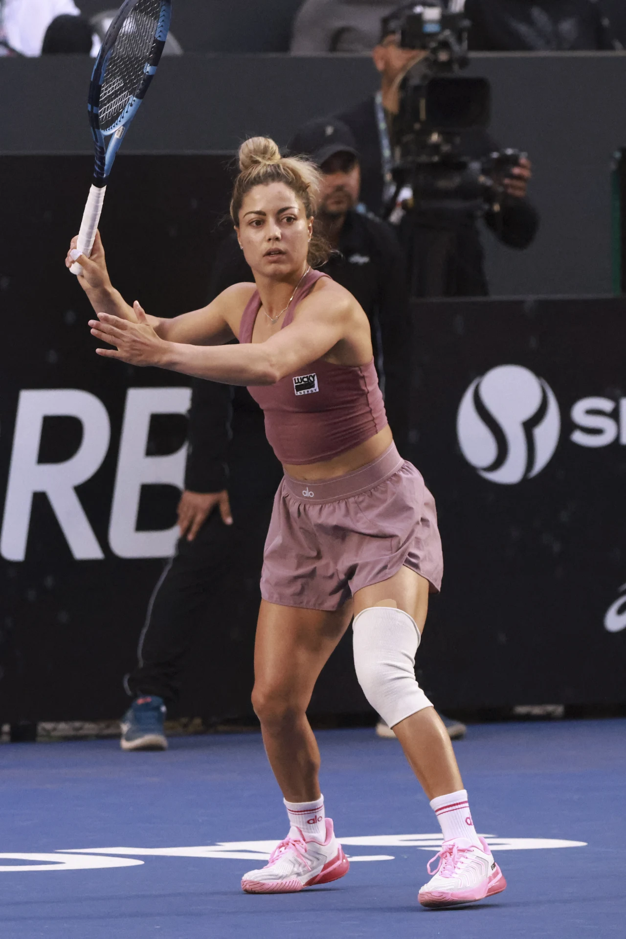Tenisistka przygotowana do odbioru piłki na korcie, ubrana w sportowy strój w odcieniach różu, z opaską na kolanie, z rakietą uniesioną w obu rękach. W tle widać fragmenty trybun oraz kamerzystę.
