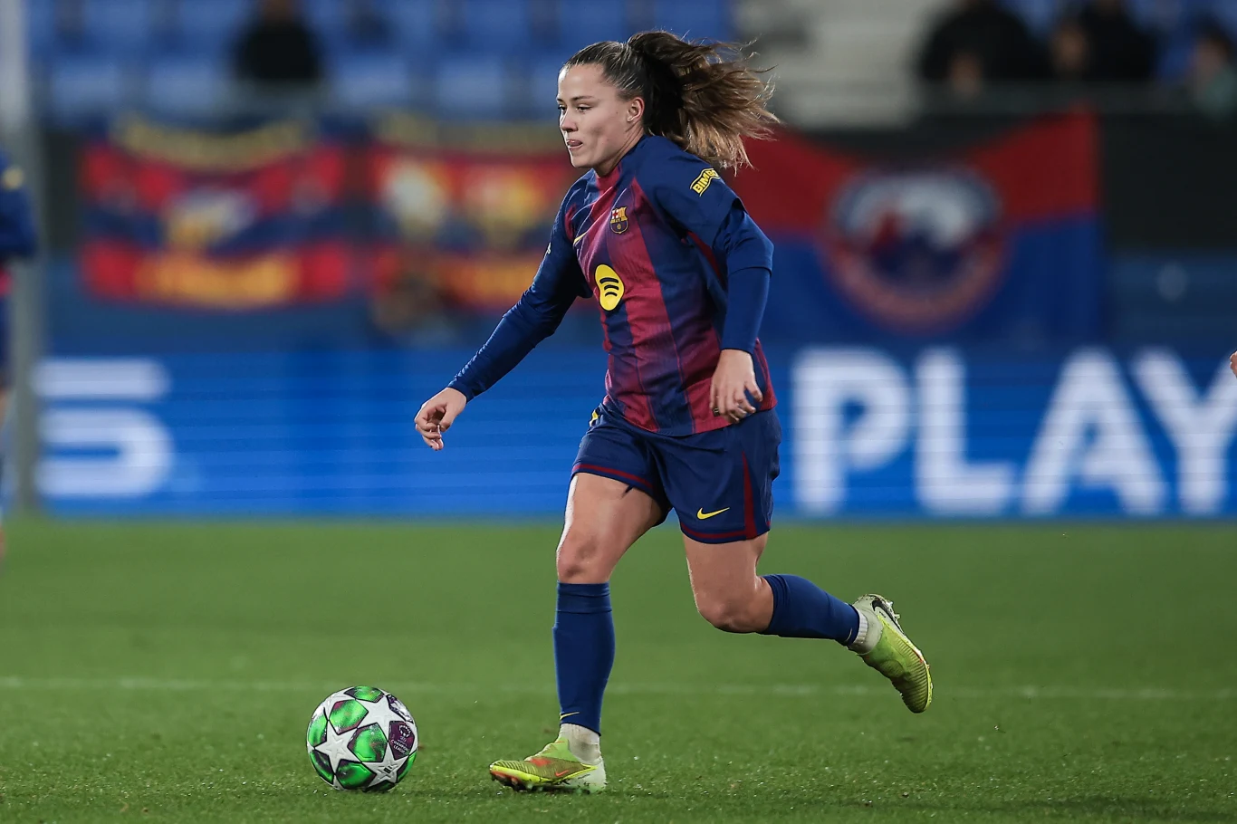 Piłkarka w stroju klubowym biegnie z piłką po zielonej murawie stadionu, w tle widoczne rozmyte flagi kibiców.