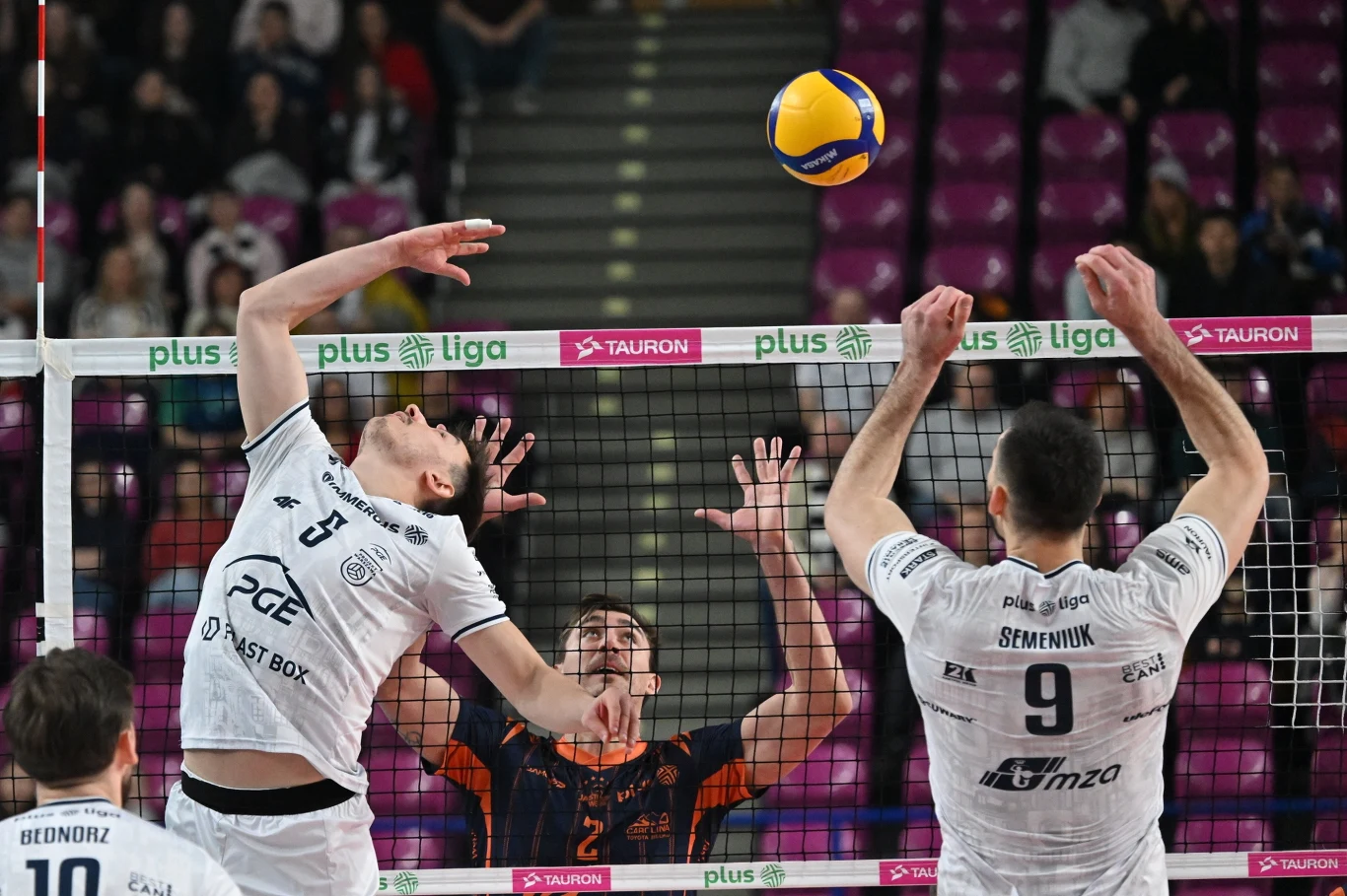 Dwóch siatkarzy w białych strojach walczy o piłkę nad siatką z zawodnikiem drużyny przeciwnej w pomarańczowo-czarnym stroju, na tle trybun z kibicami.