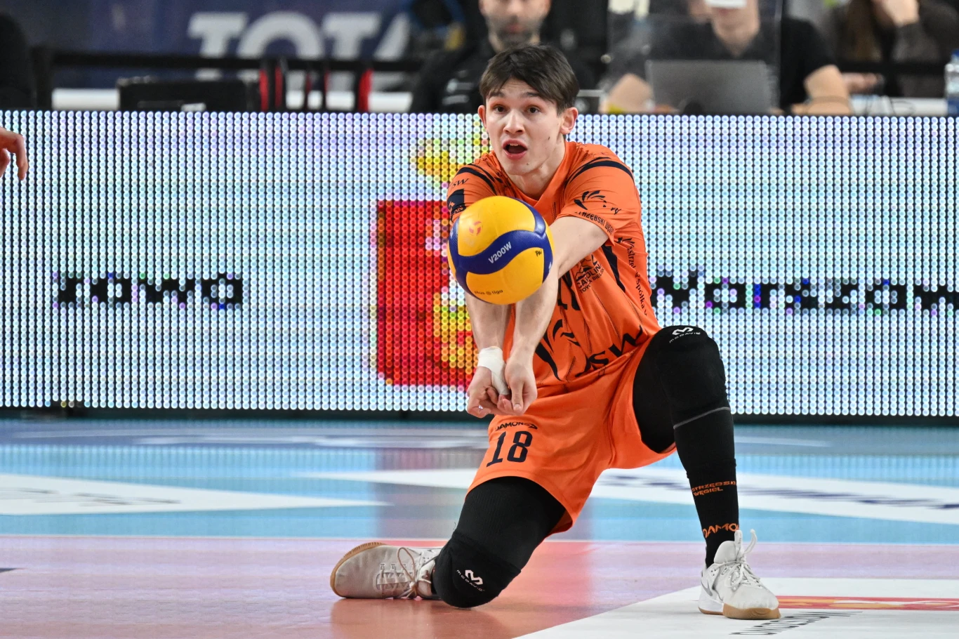 Siatkarz ubrany w pomarańczowy strój numer 18 wykonuje odbiór piłki podczas meczu na hali sportowej. Na pierwszym planie wyraźnie widać skupienie zawodnika, a tło wypełnia elektroniczna tablica informacyjna.