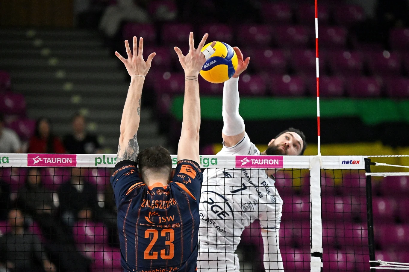 Dwóch siatkarzy podczas akcji przy siatce, jeden wykonuje atak piłką, drugi próbuje blokować, obaj skoncentrowani na grze, w tle trybuny z widocznymi kibicami.