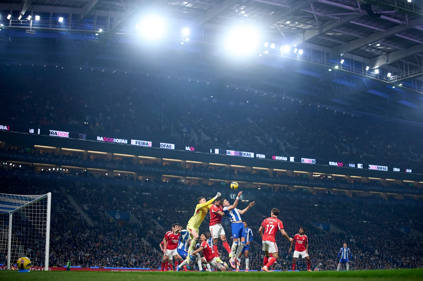 Mecz piłki nożnej na dużym stadionie, zawodnicy w czerwonych i niebieskich strojach walczą o piłkę w polu karnym, wokół widoczny tłum kibiców i silne oświetlenie nad boiskiem.