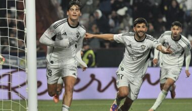 Zawodnik w niebieskim stroju drybluje piłkę, próbując minąć przeciwników w białych koszulkach. W tle widoczni są kolejni piłkarze oraz sędzia, a trybuny wypełnione są kibicami.