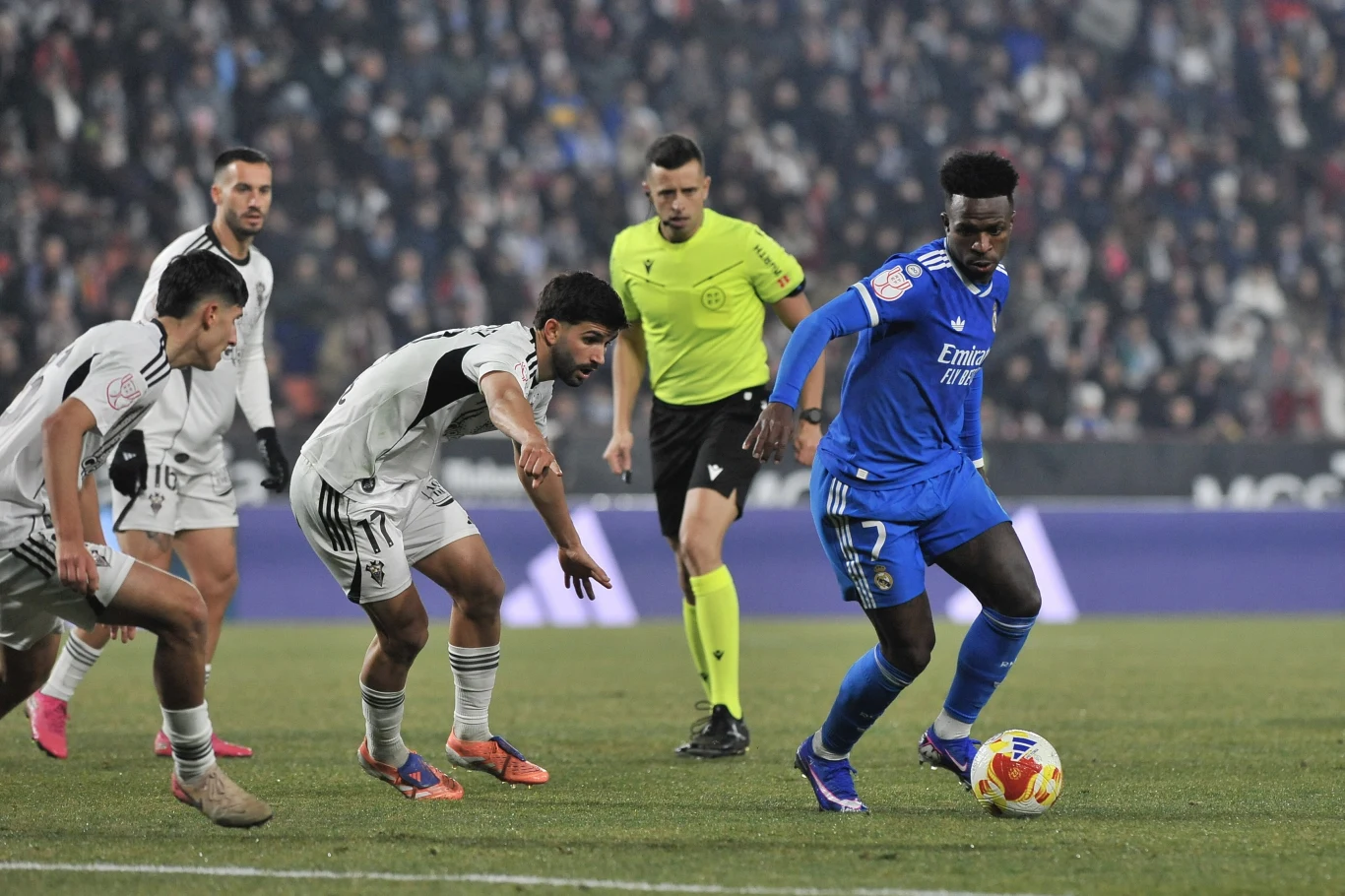 Zawodnik w niebieskim stroju drybluje piłkę, próbując minąć przeciwników w białych koszulkach. W tle widoczni są kolejni piłkarze oraz sędzia, a trybuny wypełnione są kibicami.