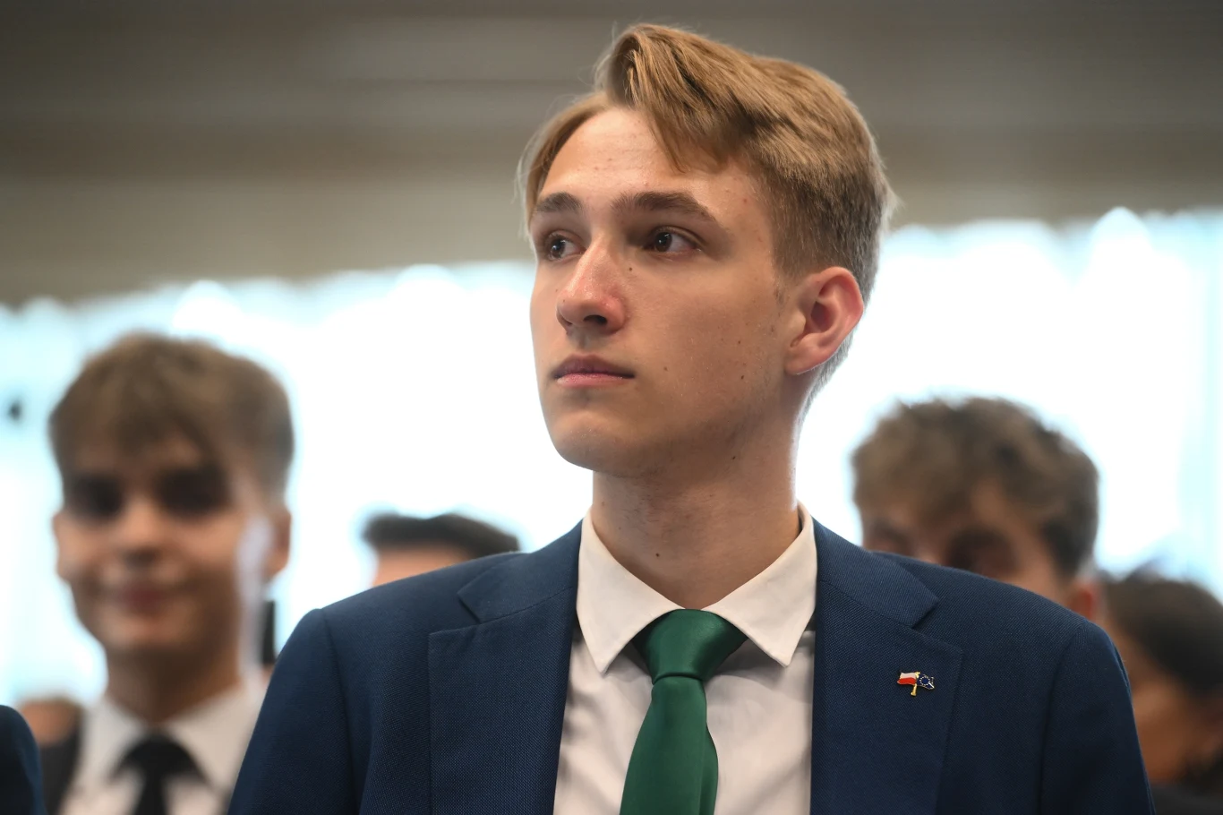 Młody mężczyzna w eleganckim garniturze z zielonym krawatem, spoglądający poważnie przed siebie, w tle kilku innych młodych mężczyzn o rozmytych twarzach.