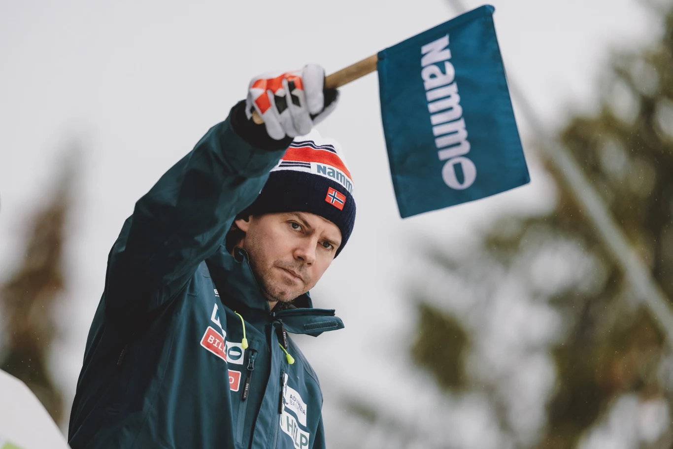 Mężczyzna w kurtce z naszywkami sponsorów i czapce z norweską flagą trzyma niebieską flagę z napisem Nammo, w tle nieostre drzewa i zimowa sceneria.